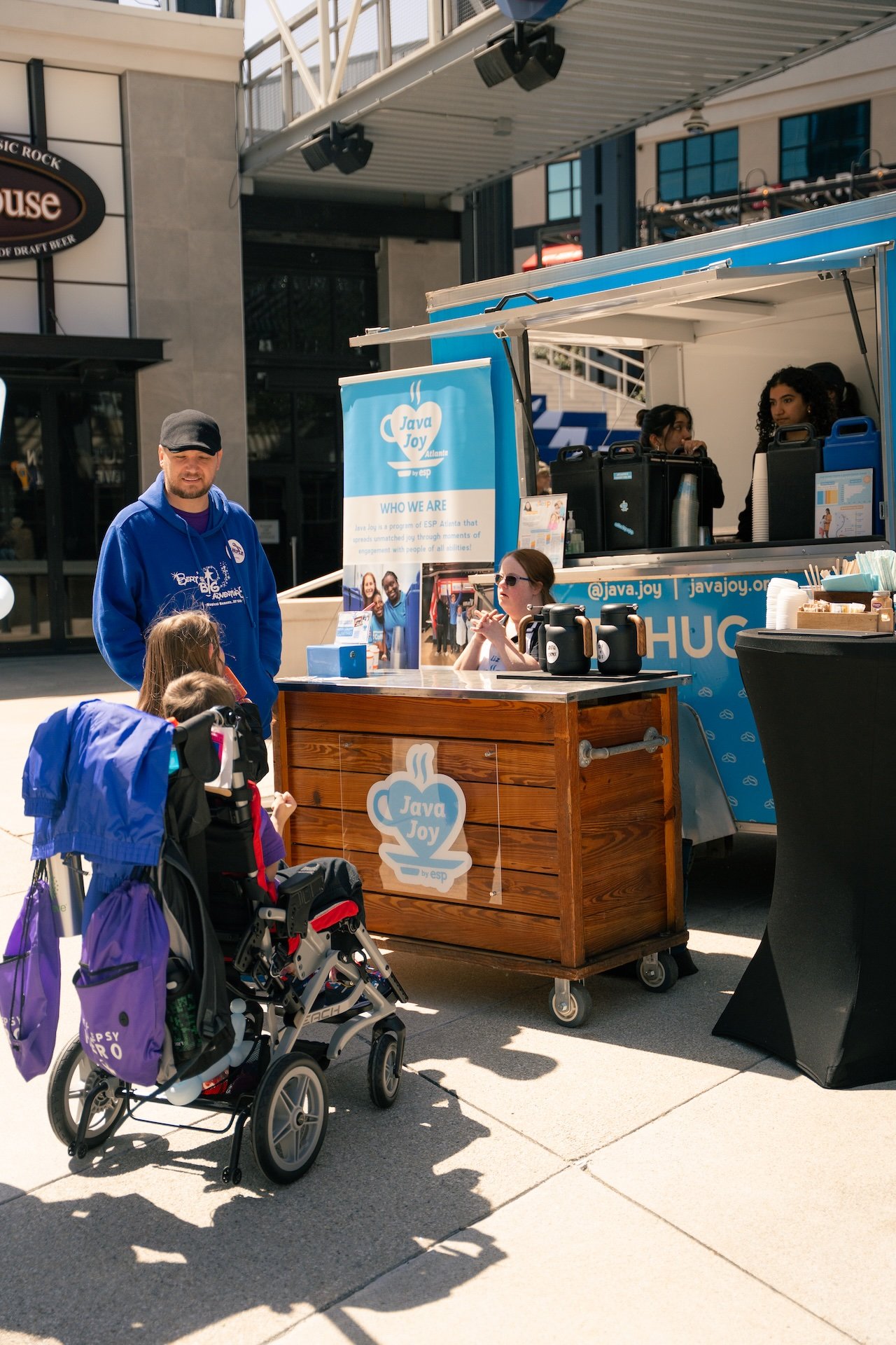 A man and two young girls at a mobile booth for Java Joy, an organization supporting people of all abilities. The booth has a sign with the organization's logo and information, and there are coffee dispensers on the counter. The man is wearing a blue