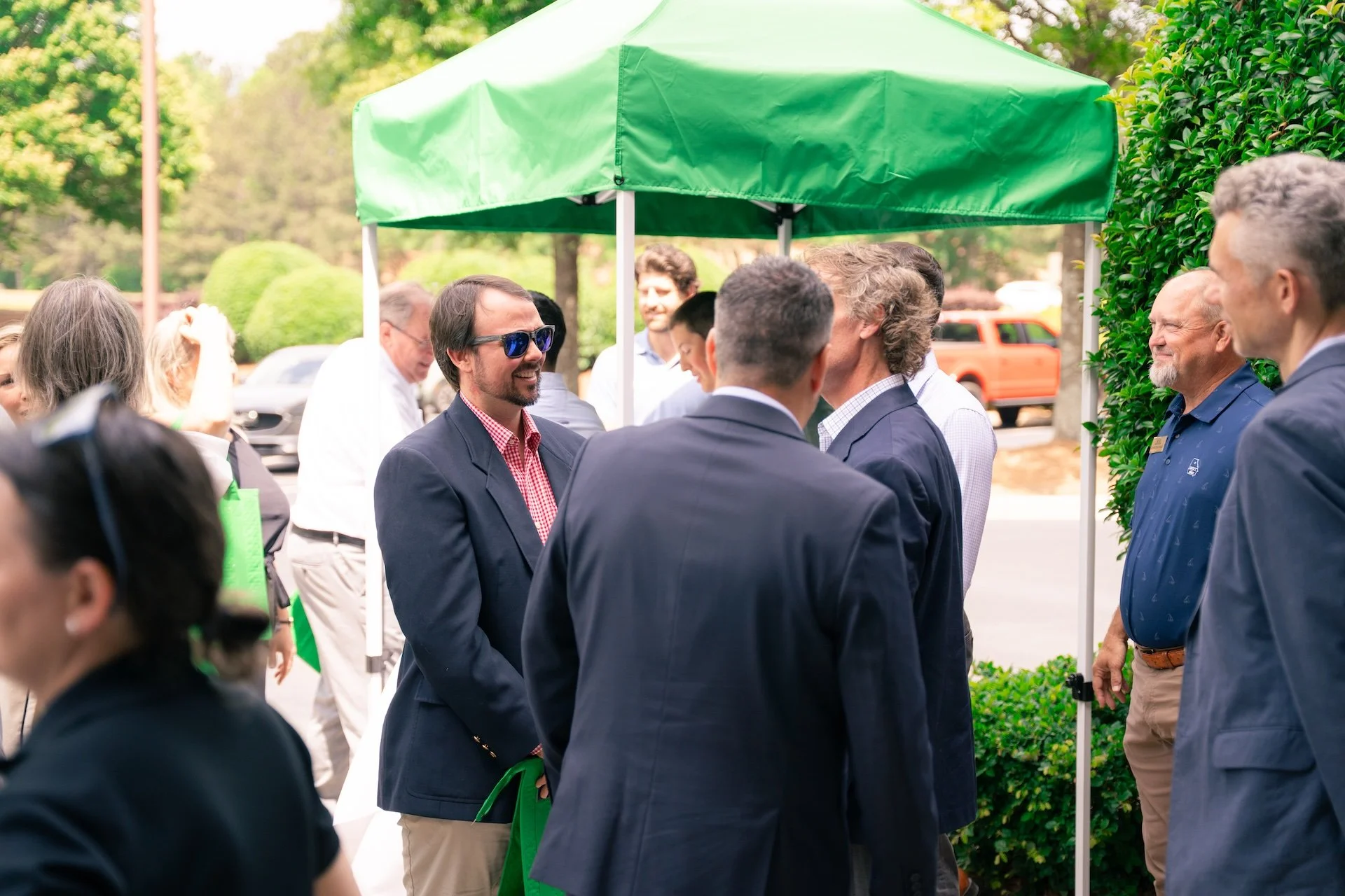 Group of people gathered outdoors under a green canopy, engaged in conversation, with trees and cars in the background.