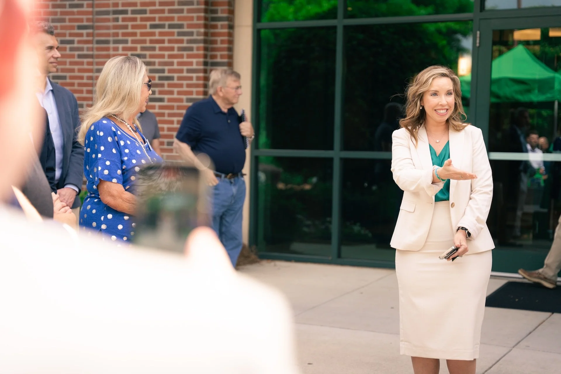 A woman in a white suit is speaking to a group of people outside a building with green-tinted glass windows. She is smiling, holding a phone, and gesturing with her hand.