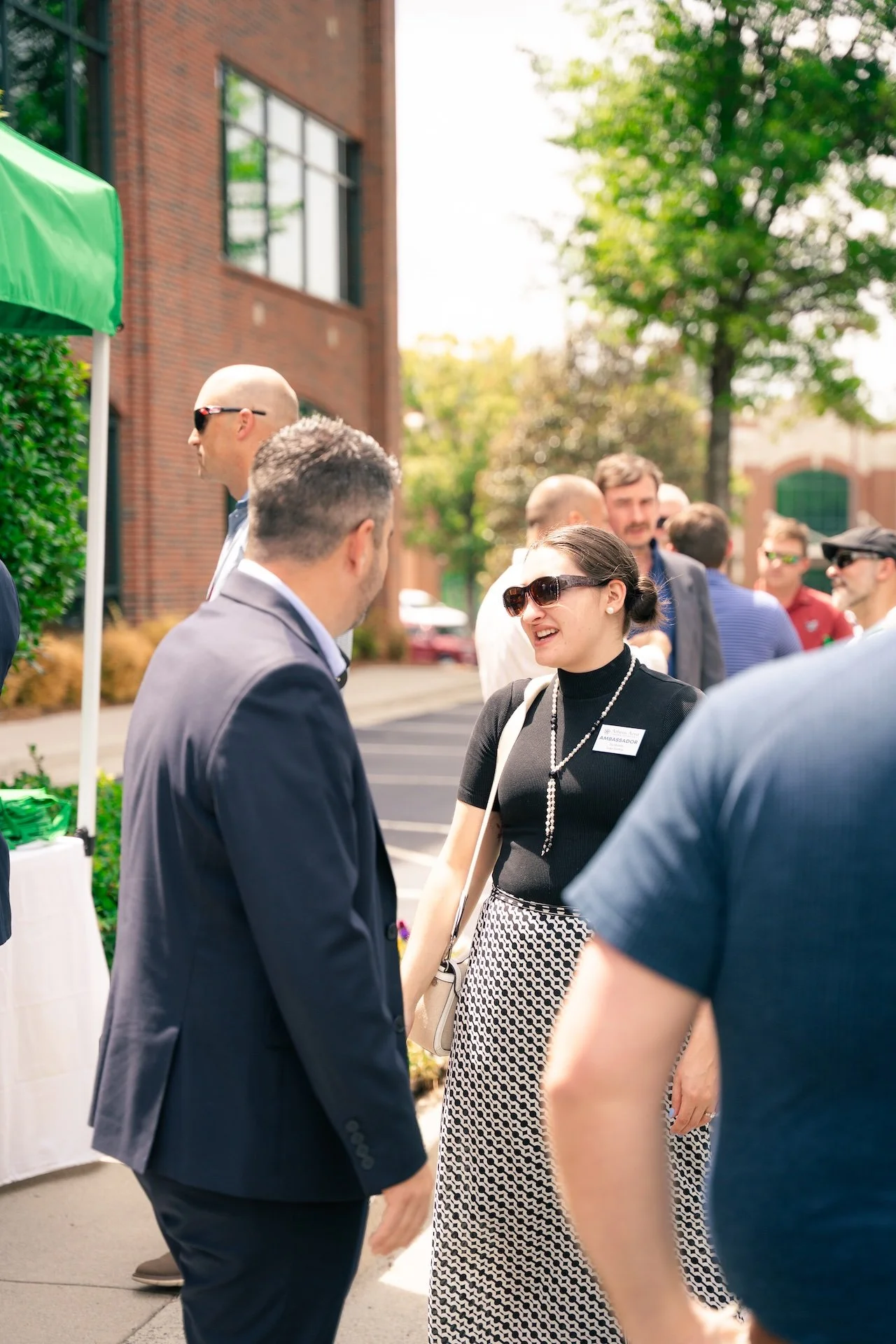 People socializing outdoors at a professional event, with a woman in black top and patterned skirt smiling and talking to a man in a suit.