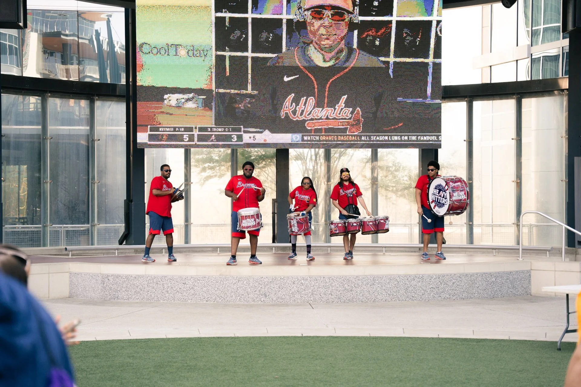 A musical band wearing red shirts and blue shorts playing drums on an outdoor stage with a large digital screen behind them. The screen displays a person in Atlanta Braves attire and some text related to baseball.