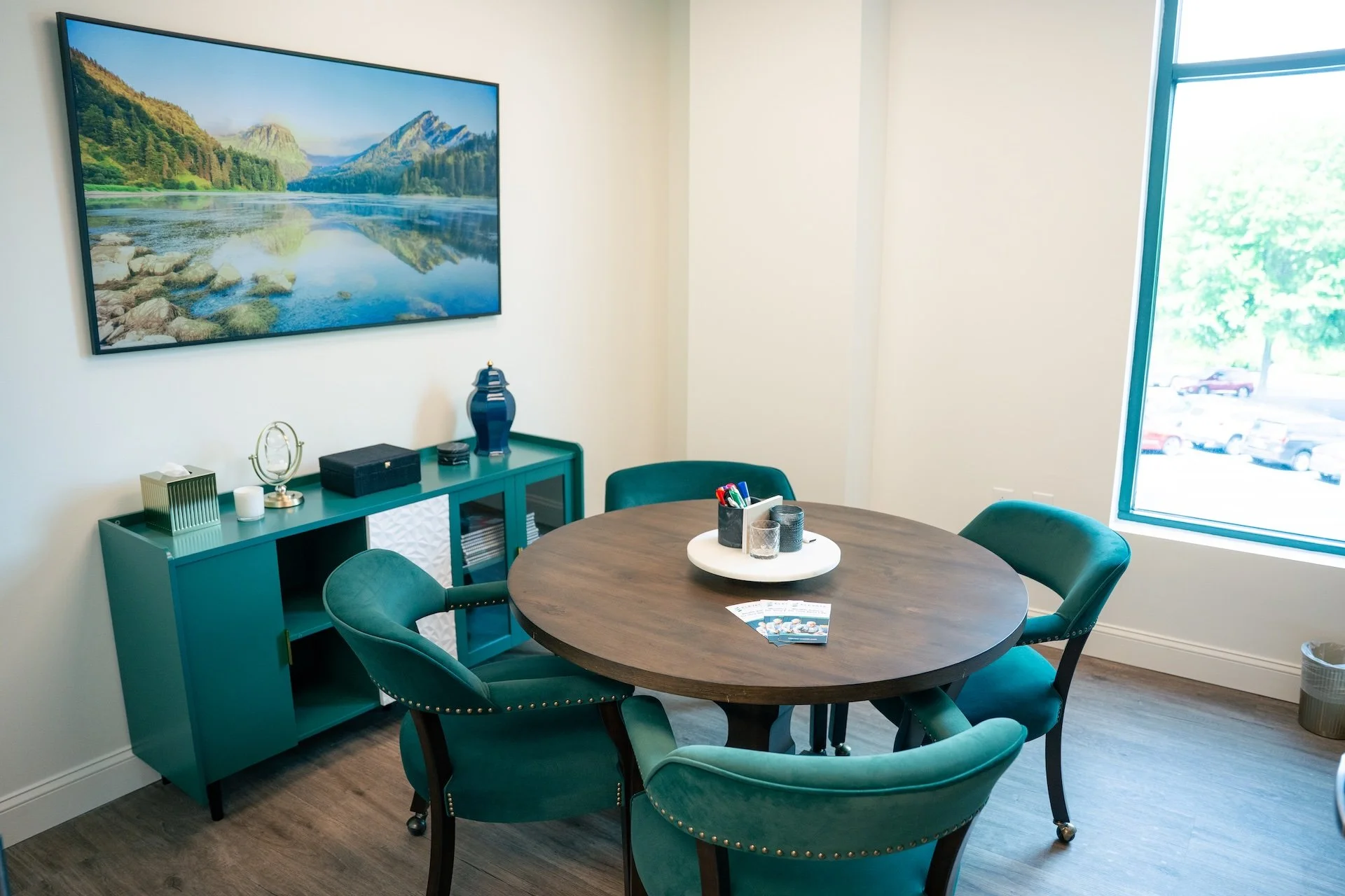 A modern conference room with a round wooden table encircled by teal upholstered chairs, a teal sideboard with decorative objects and books, a large wall-mounted landscape painting, a window showing parked cars outside, and a tray with pens, candles,
