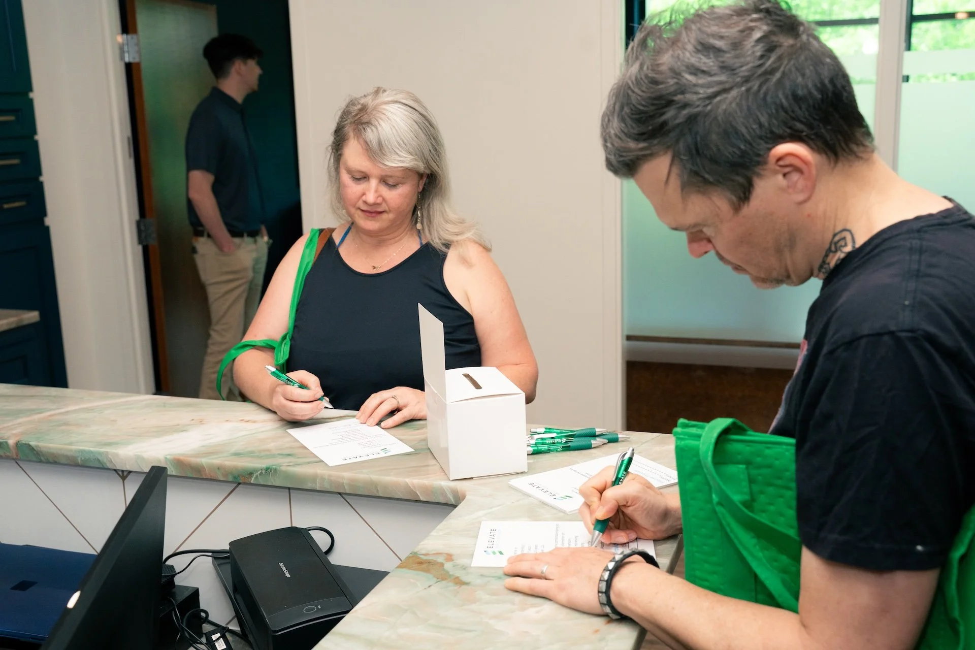 A woman and a man signing documents at a registration or check-in counter, with a woman in the background near a door.