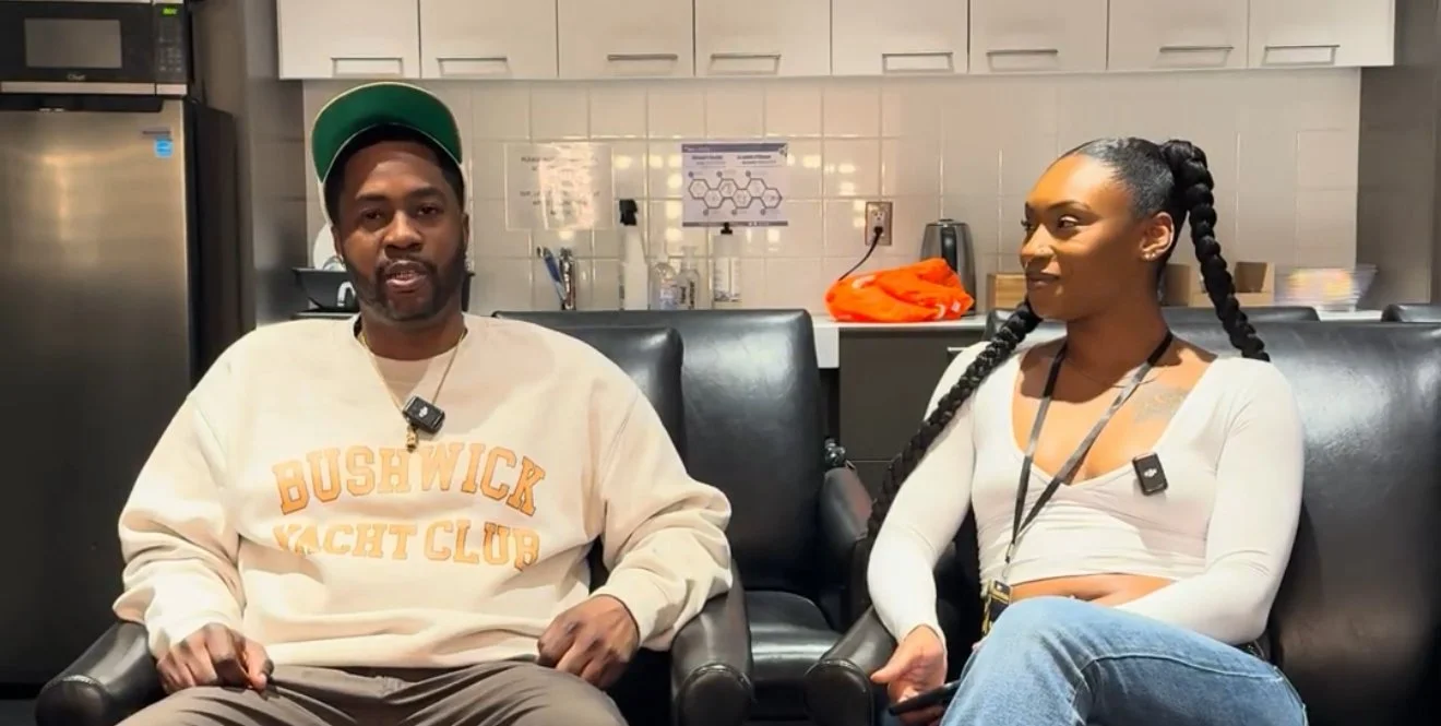 A man and woman sitting on black chairs in a room with a kitchen background, wearing microphones around their necks, engaged in a conversation.