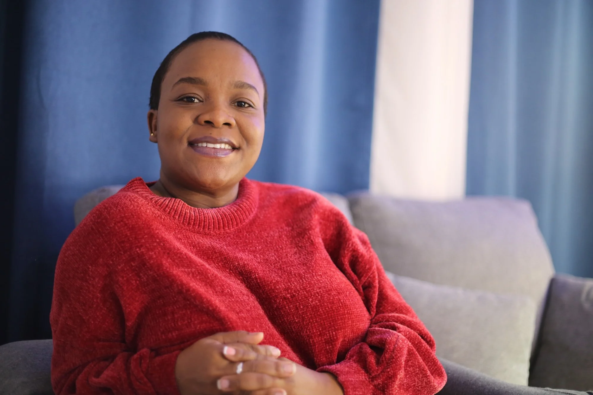 A woman sitting on a gray sofa, smiling, with blue curtains in the background, wearing a red sweater.