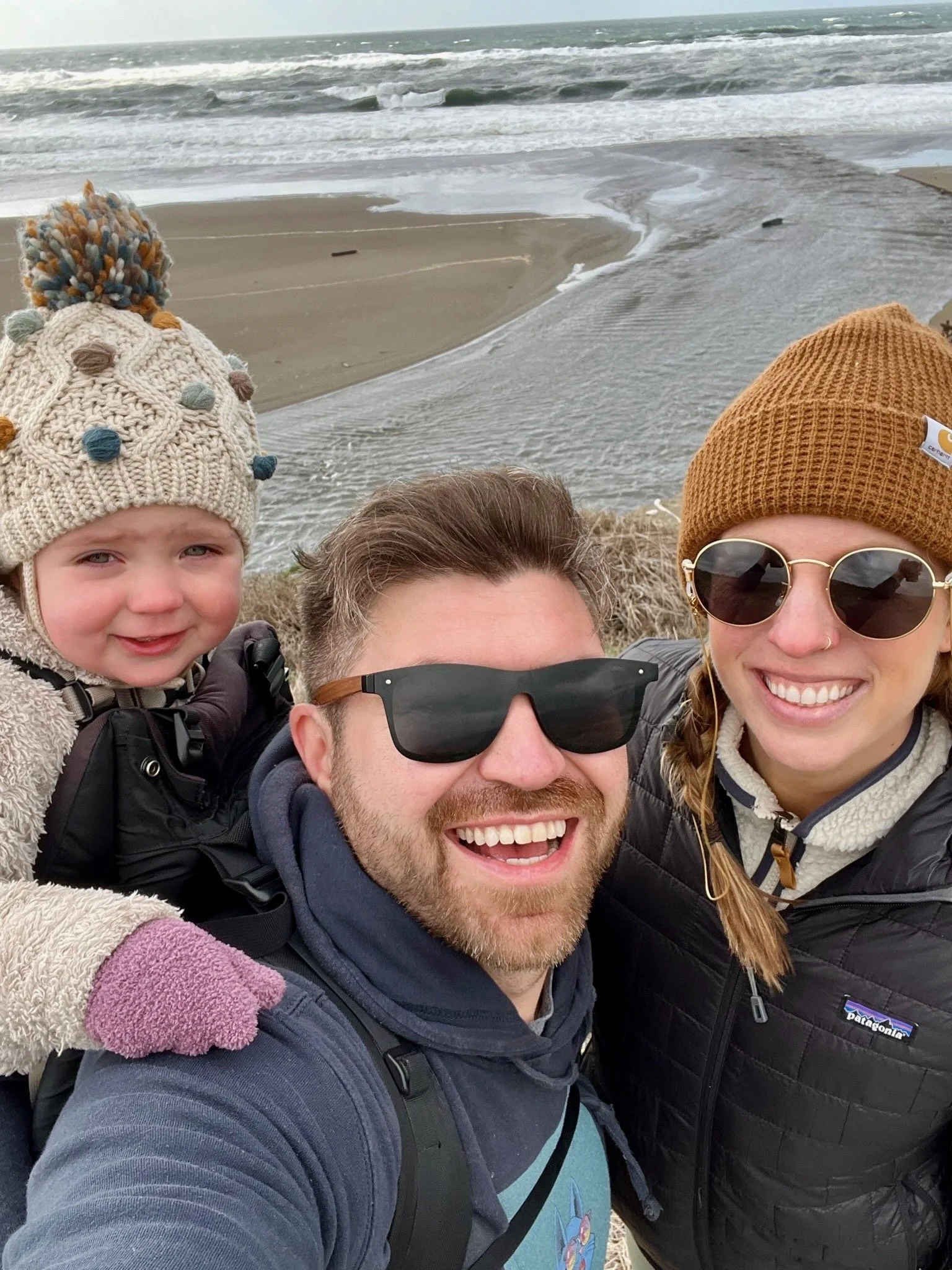 A smiling family of three on the beach, with the ocean and waves in the background. The man in the center wears sunglasses, the woman on the right wears sunglasses and a brown beanie, and the young girl on the left wears a colorful knit hat with pom-poms.