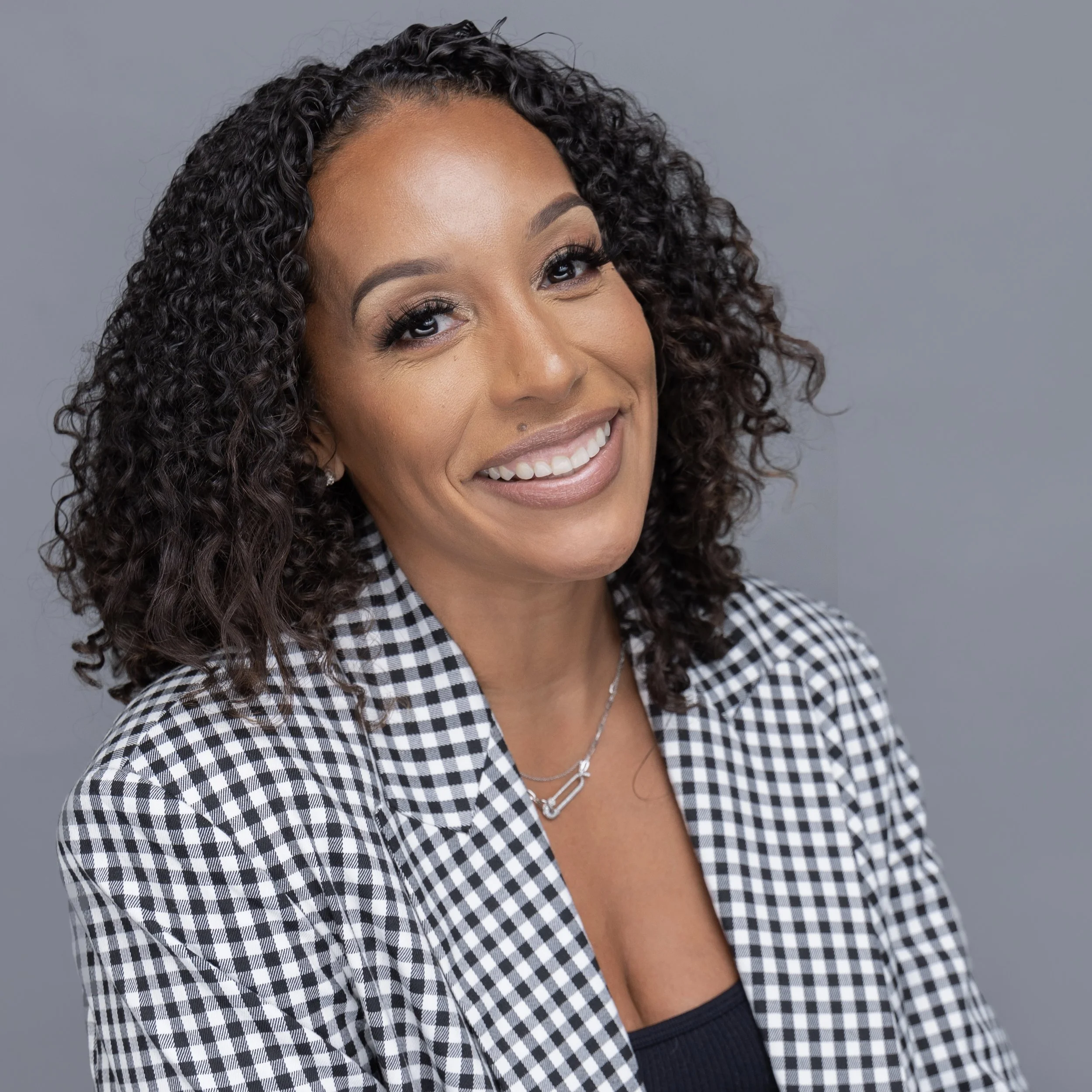 Close-up portrait of a smiling woman with curly dark hair, wearing a black and white checkered blazer and a black top, against a plain gray background.