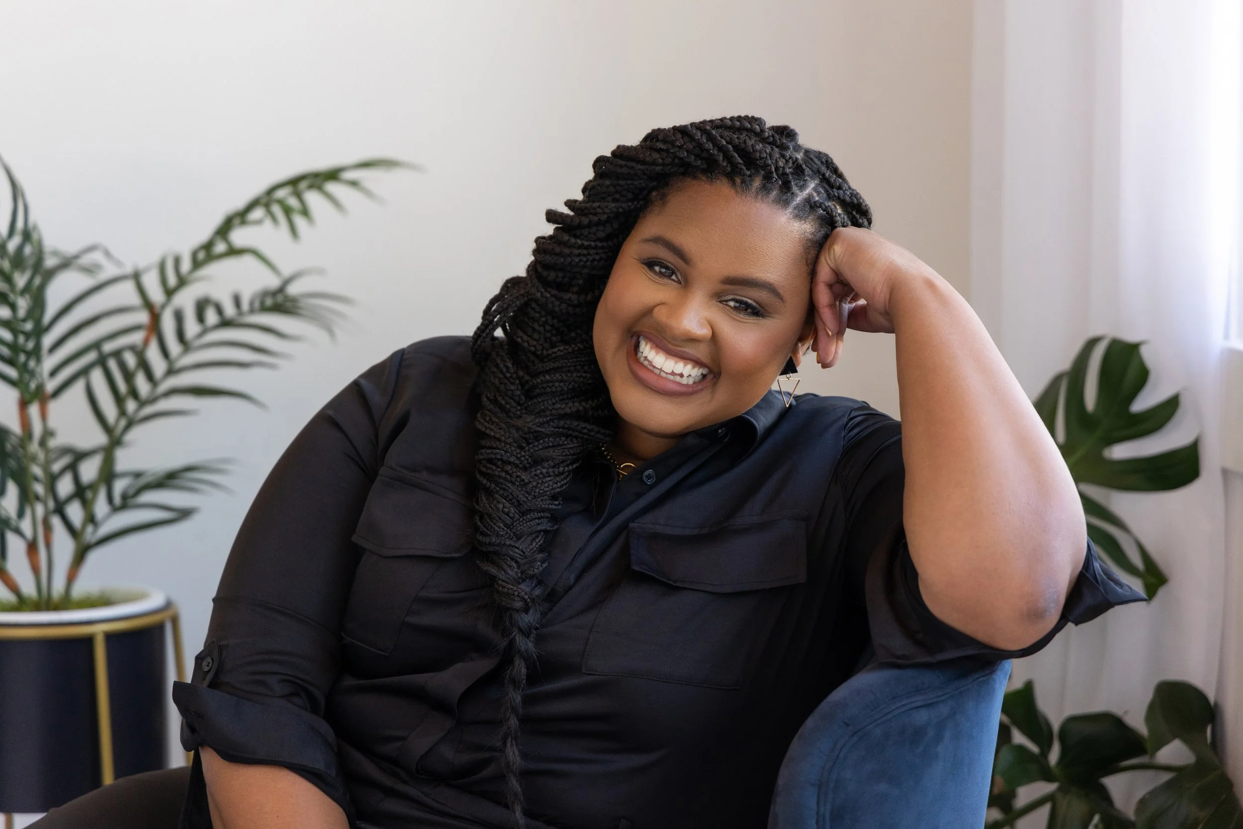 A woman with braided hair smiling, sitting in a room with houseplants and natural light.