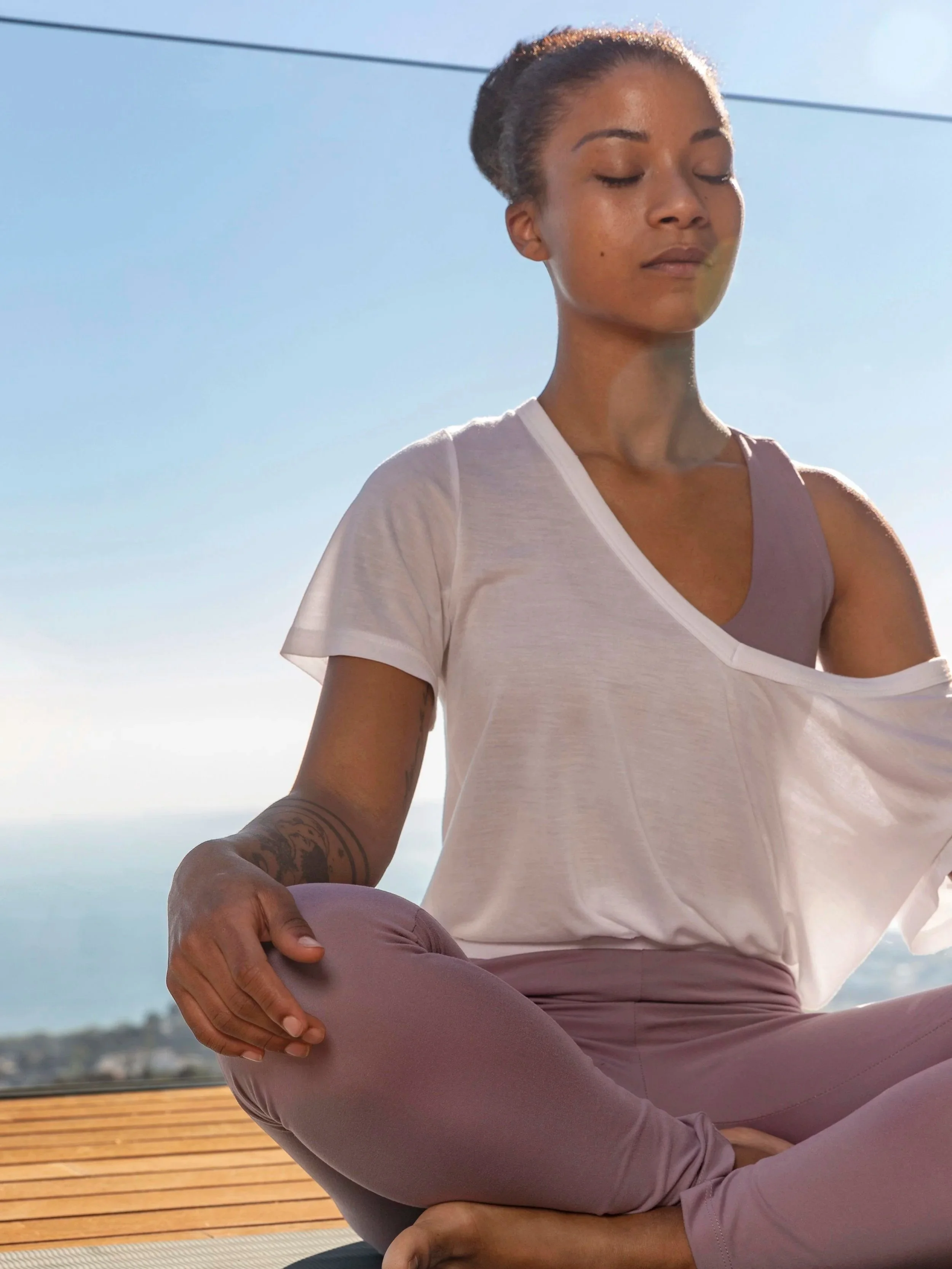 Woman meditating outdoors on a yoga mat with ocean view, wearing a white t-shirt and mauve leggings, sitting cross-legged with eyes closed.