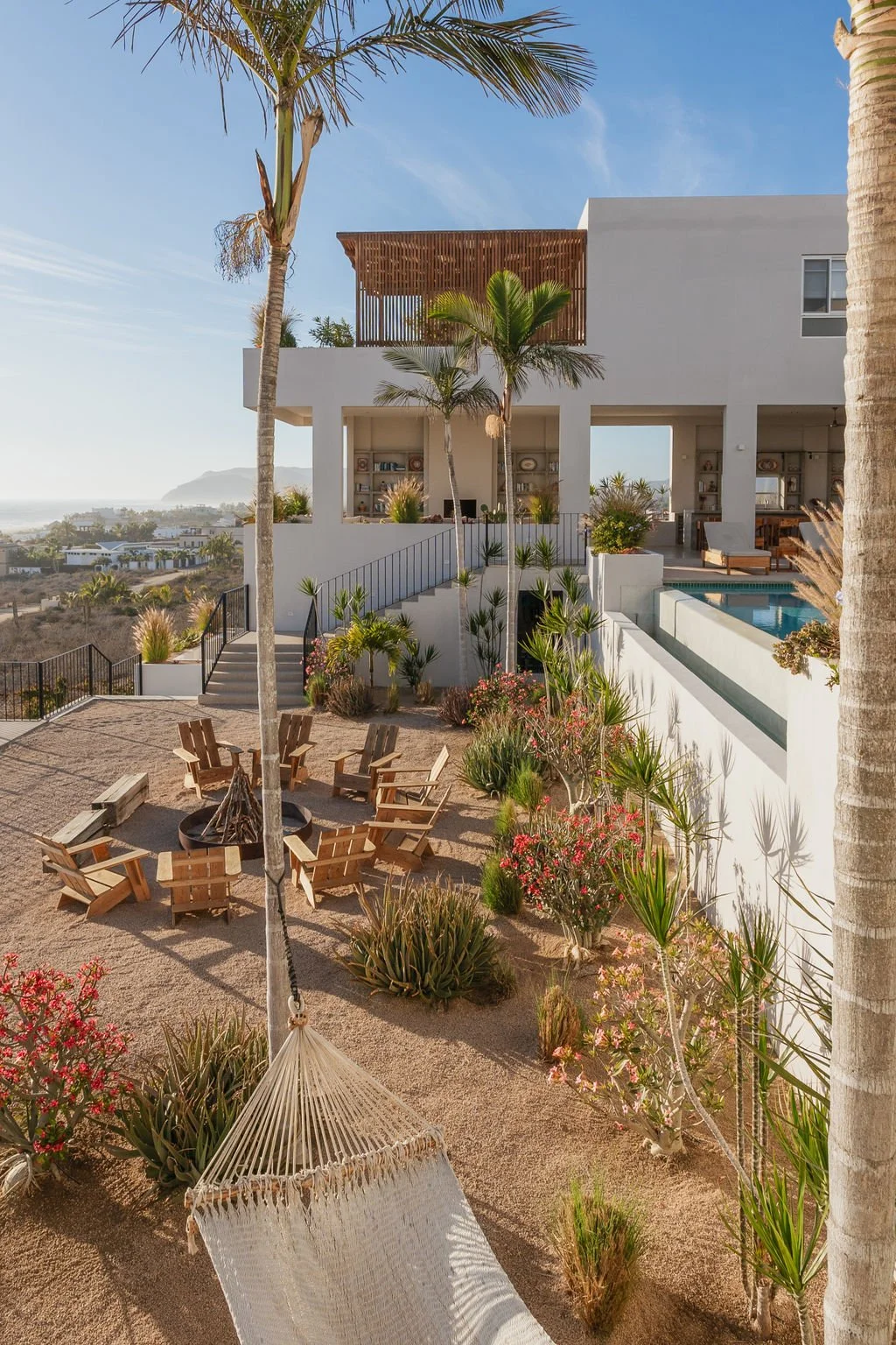 Modern white house with outdoor patio, swimming pool, palm trees, and sandy yard with wooden chairs and a hammock, overlooking a coastal landscape.