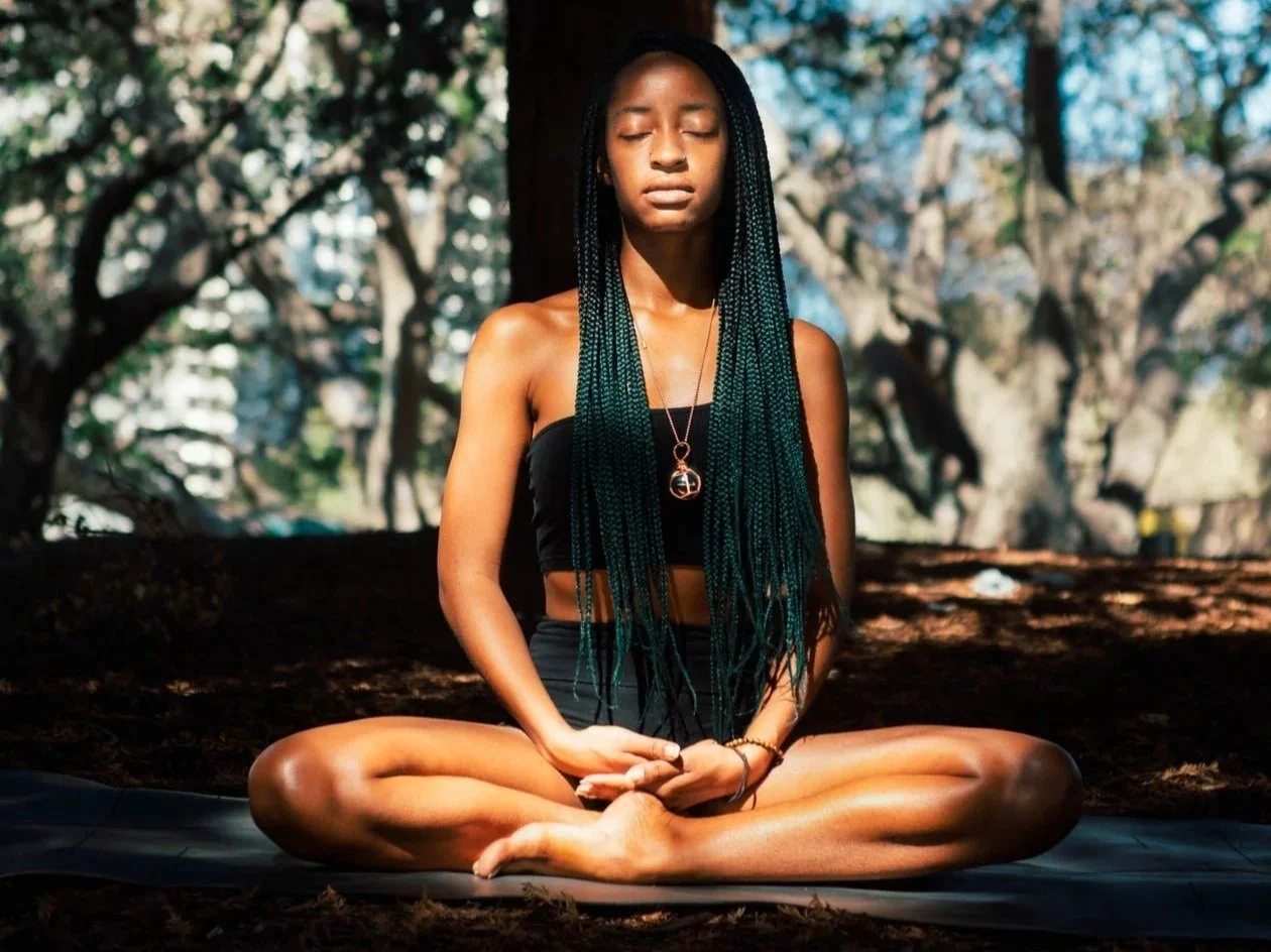 A woman with long braided hair sits cross-legged outdoors in a meditative pose with eyes closed, in a forest setting with trees and sunlight filtering through.