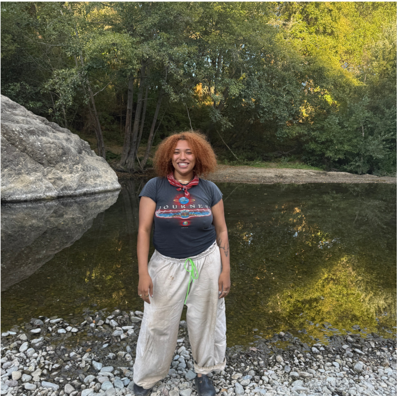 A woman with curly red hair smiling and standing on a rocky shoreline near a calm river with trees in the background.