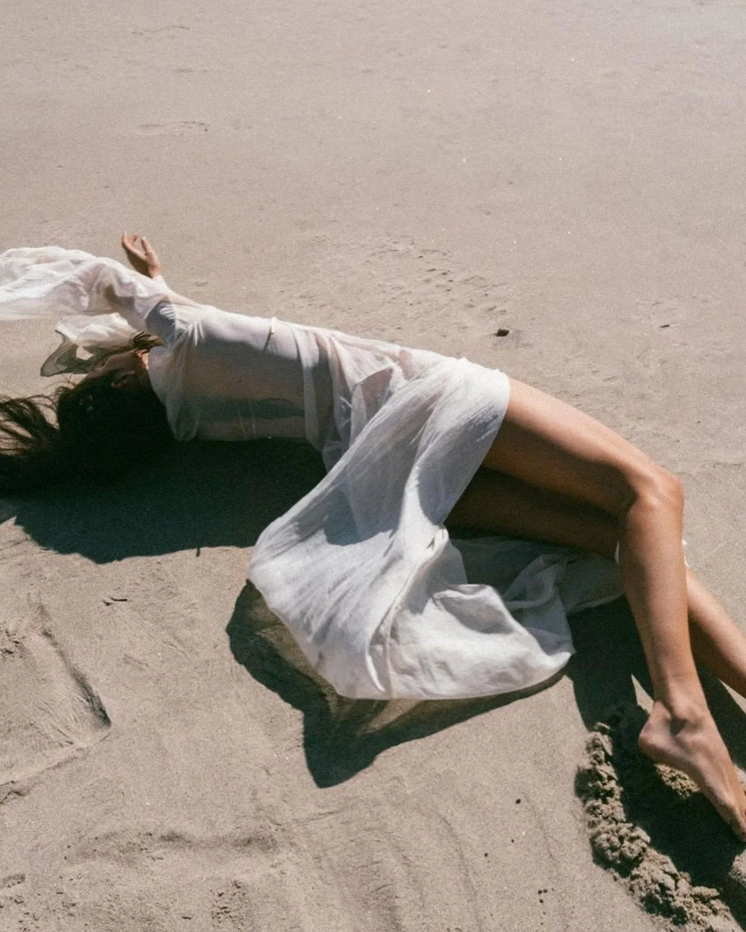 A woman lying on her side on the sandy beach, wearing a sheer white dress with dark underneath, with one arm up and her hair spread on the sand.