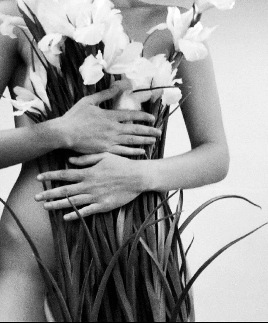 Black and white photo of a person's torso and arms holding a large bouquet of flowers, including lilies, with some tall grass or leaves at the bottom.