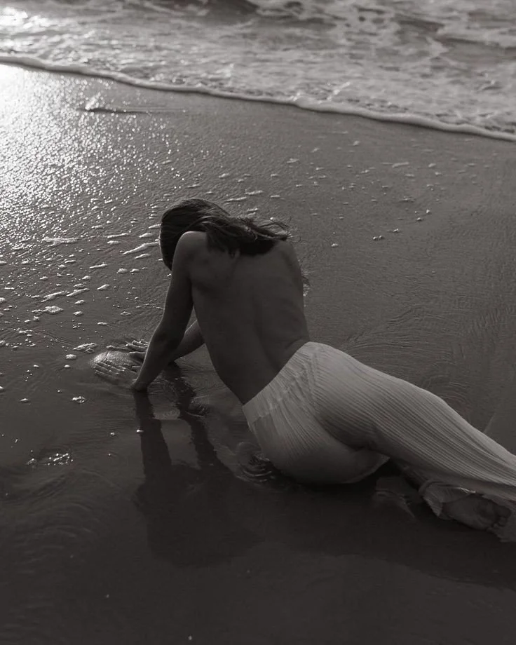 A person with long hair kneeling on the beach in shallow water, with waves approaching.