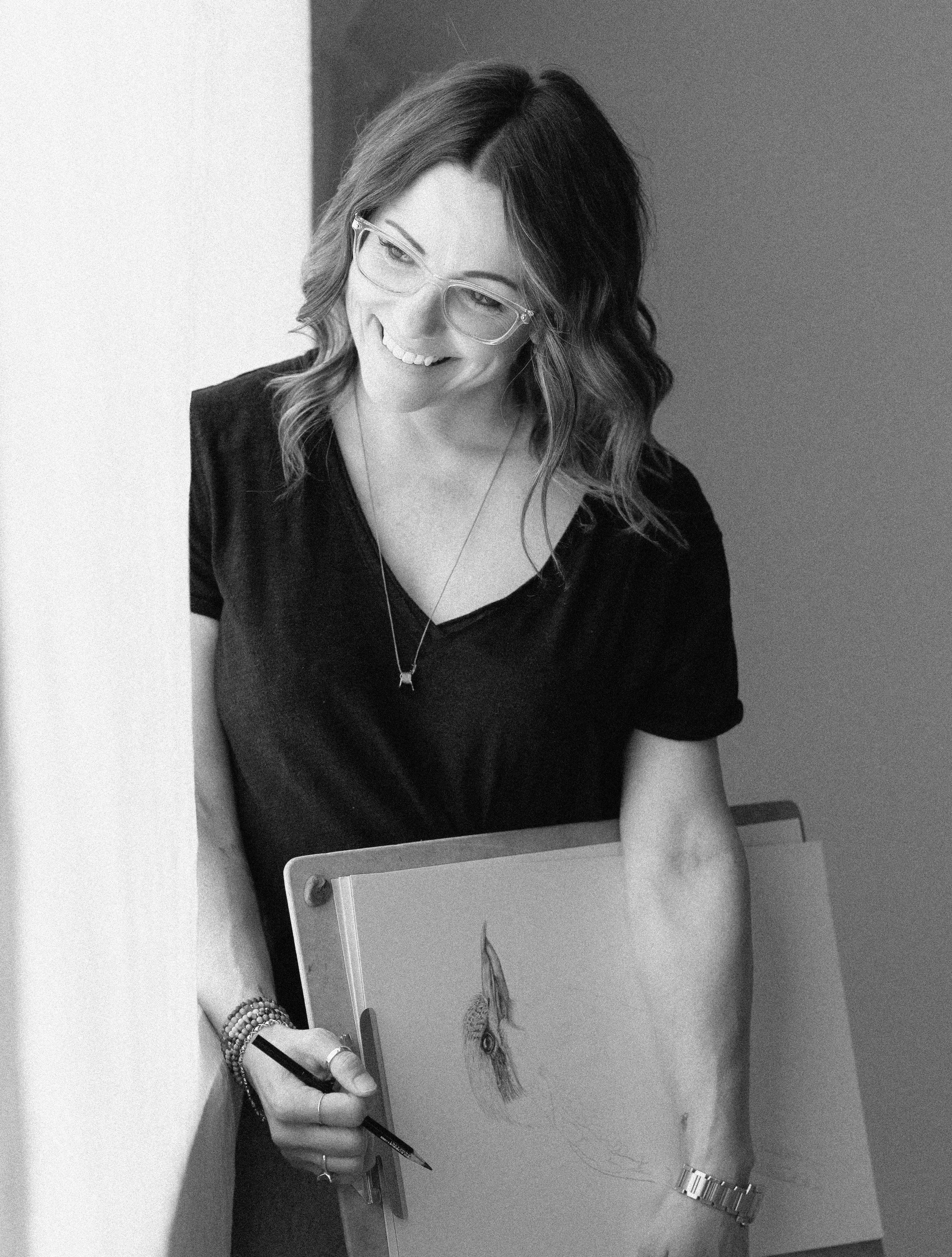 Black and white photo of a woman with wavy hair, glasses, wearing a watch and bracelets, smiling, drawing a bird on a sketchpad at a desk.