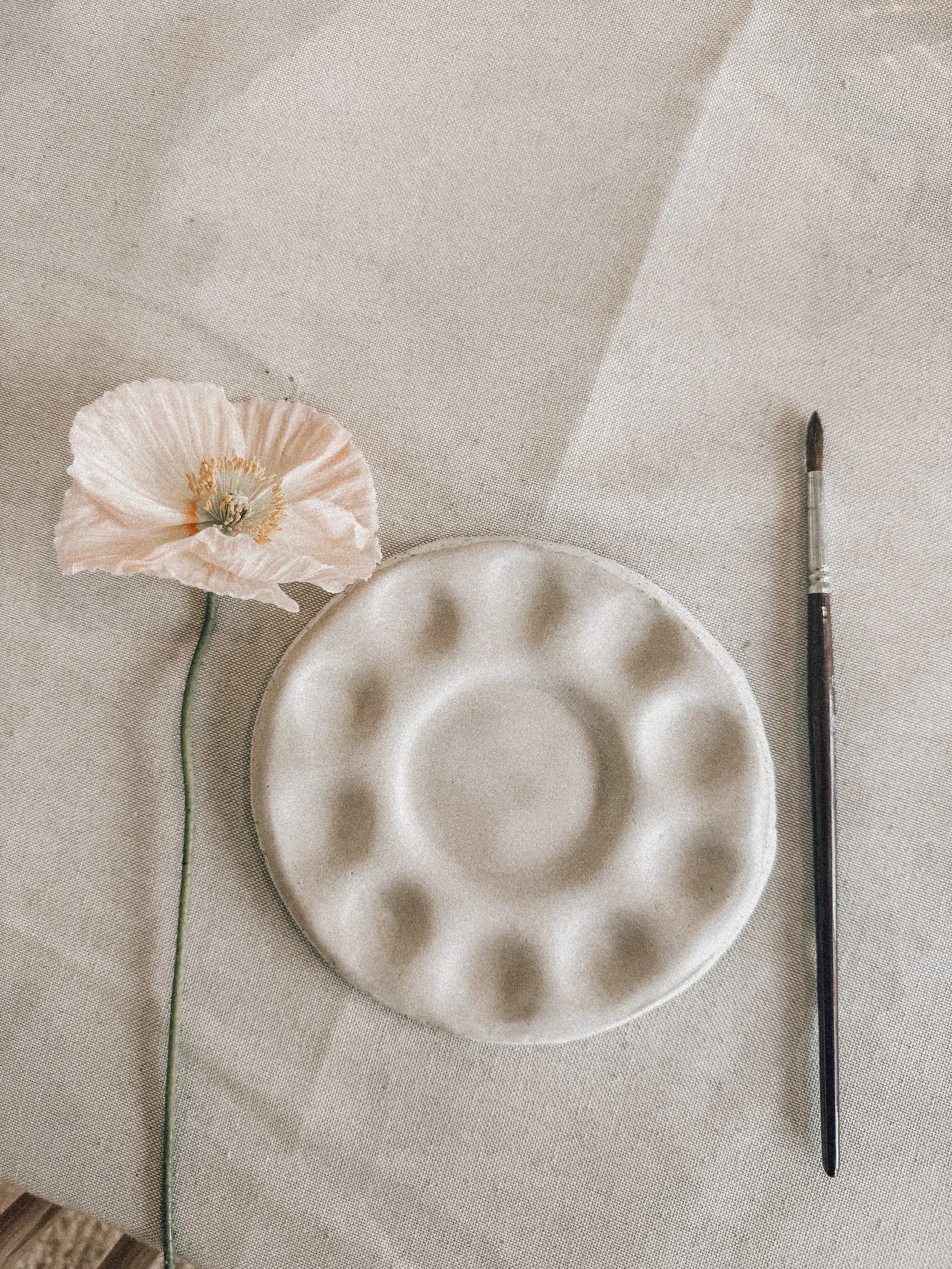 A white poppy flower with a green stem, a ceramic bowl, and a small paintbrush with a black tip on a neutral beige fabric surface.