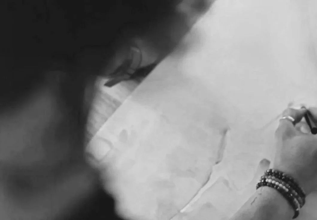 Black and white photo of a person writing on a large sheet of paper with a pen, wearing jewelry on their wrist.