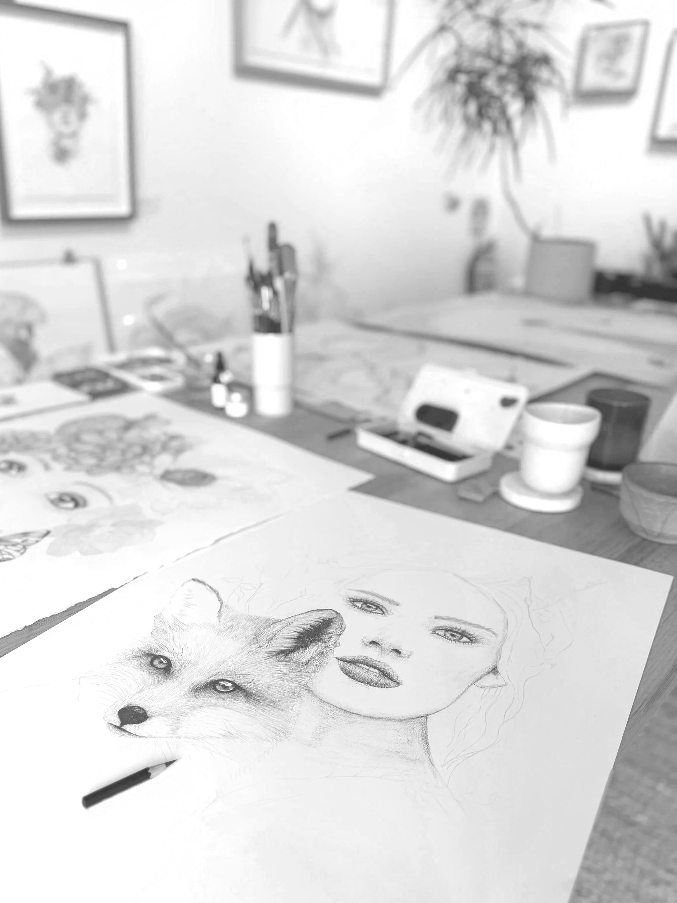 A detailed pencil drawing of a woman's face and a wolf's face on a sheet of paper, set on a table in an artist's workspace.