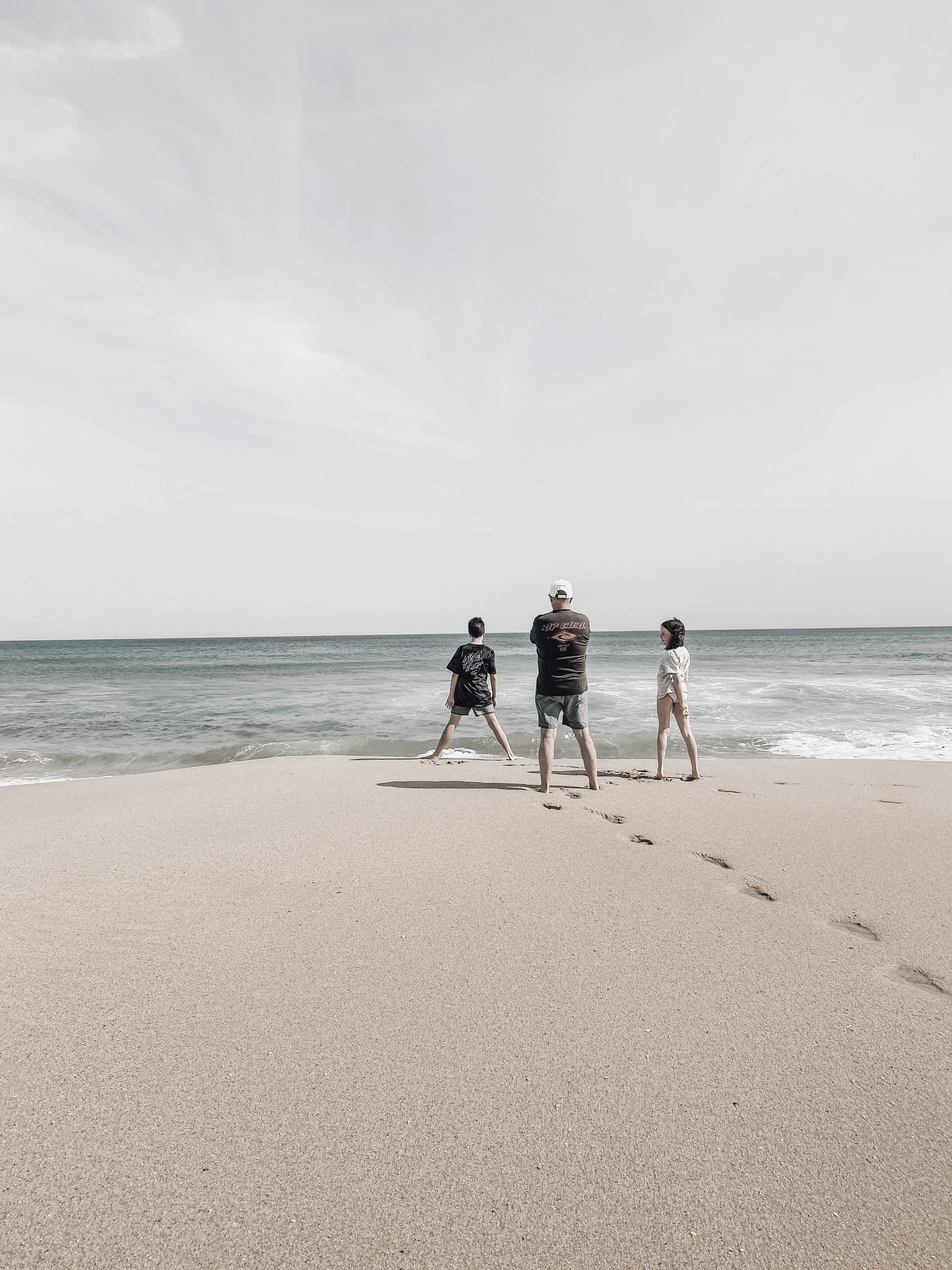 Three people, two children and an adult, standing on a sandy beach facing the ocean, with footprints trailing behind them, under a cloudy sky.