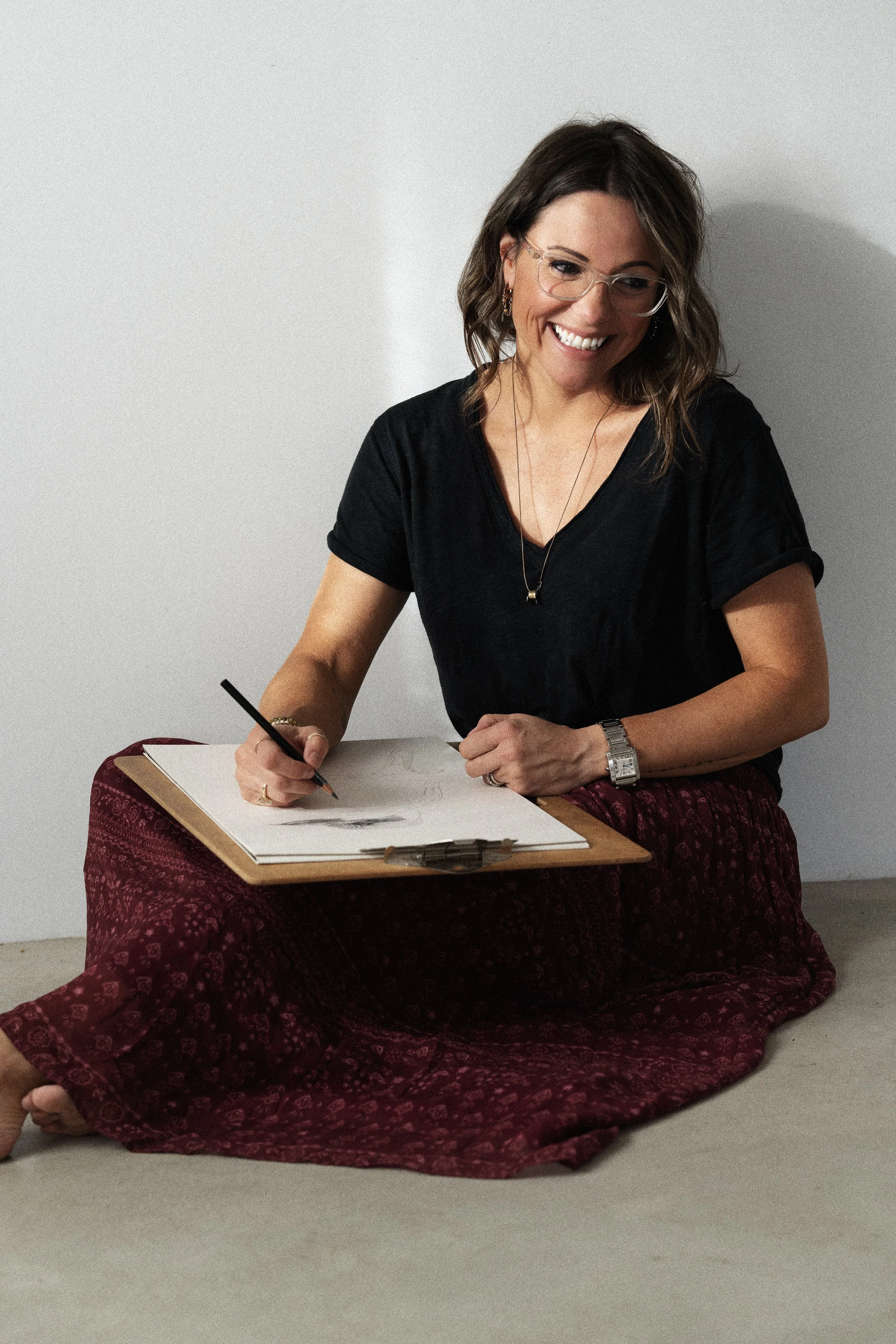 A woman sitting on the floor, wearing glasses, a black top, and a red patterned skirt, smiling while writing in a sketchbook.