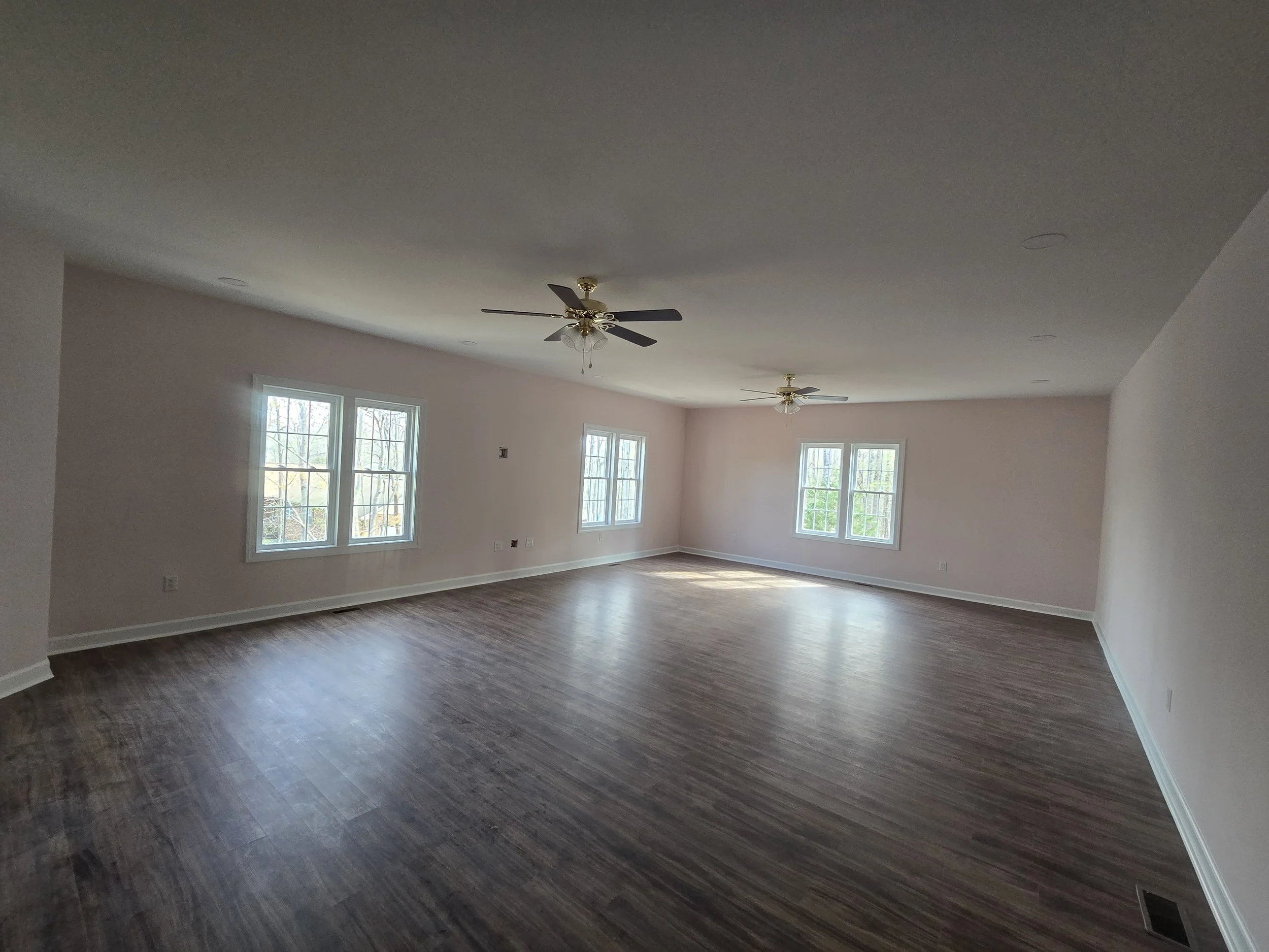 Empty living room with light-colored walls, multiple windows, and hardwood floors, with two ceiling fans.
