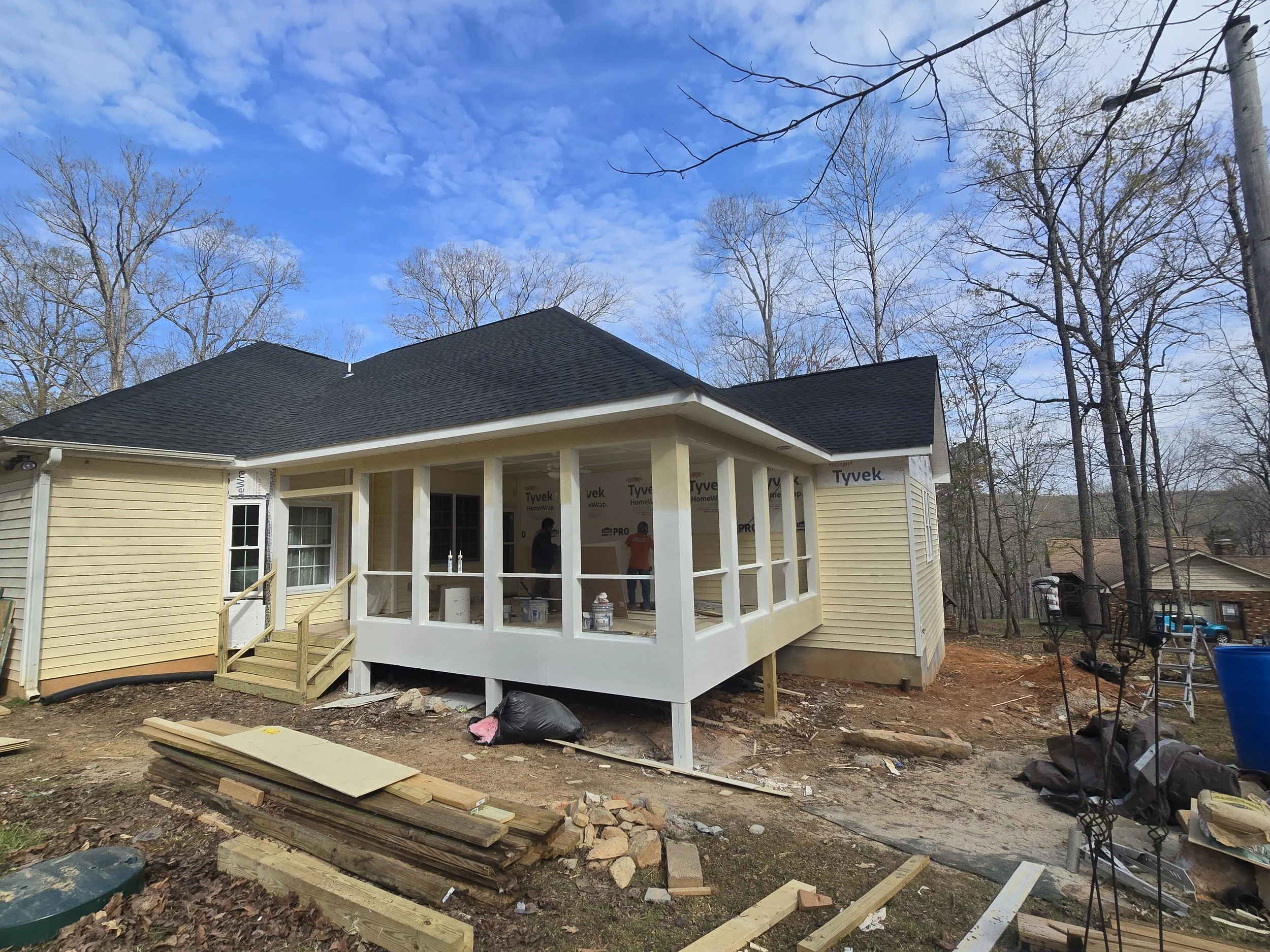 Under construction house with a screened porch, yellow siding, black roof, and blue sky with clouds, in a wooded area.