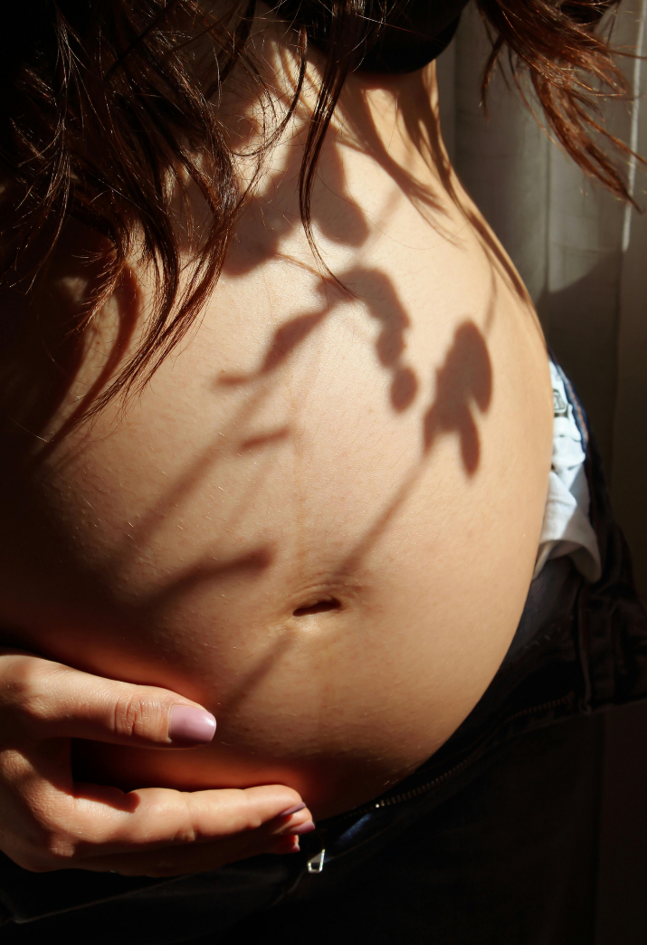 Close-up of a pregnant woman's belly with shadows of leaves and flowers cast on her skin, her hand resting below.