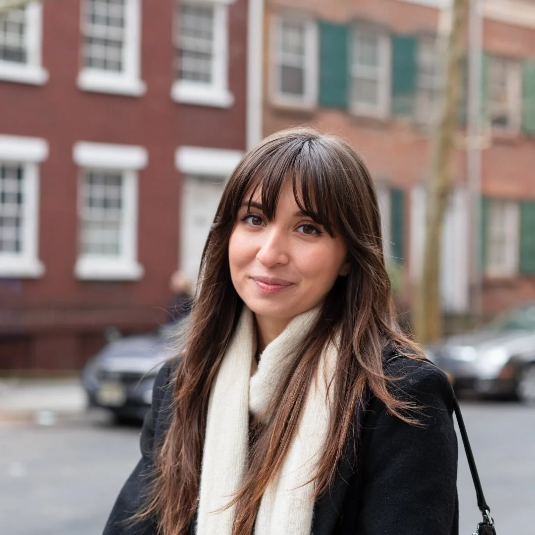Portrait of a woman with long brown hair and bangs, wearing a cream-colored scarf and black coat, standing outdoors with a city street and brick buildings in the background.