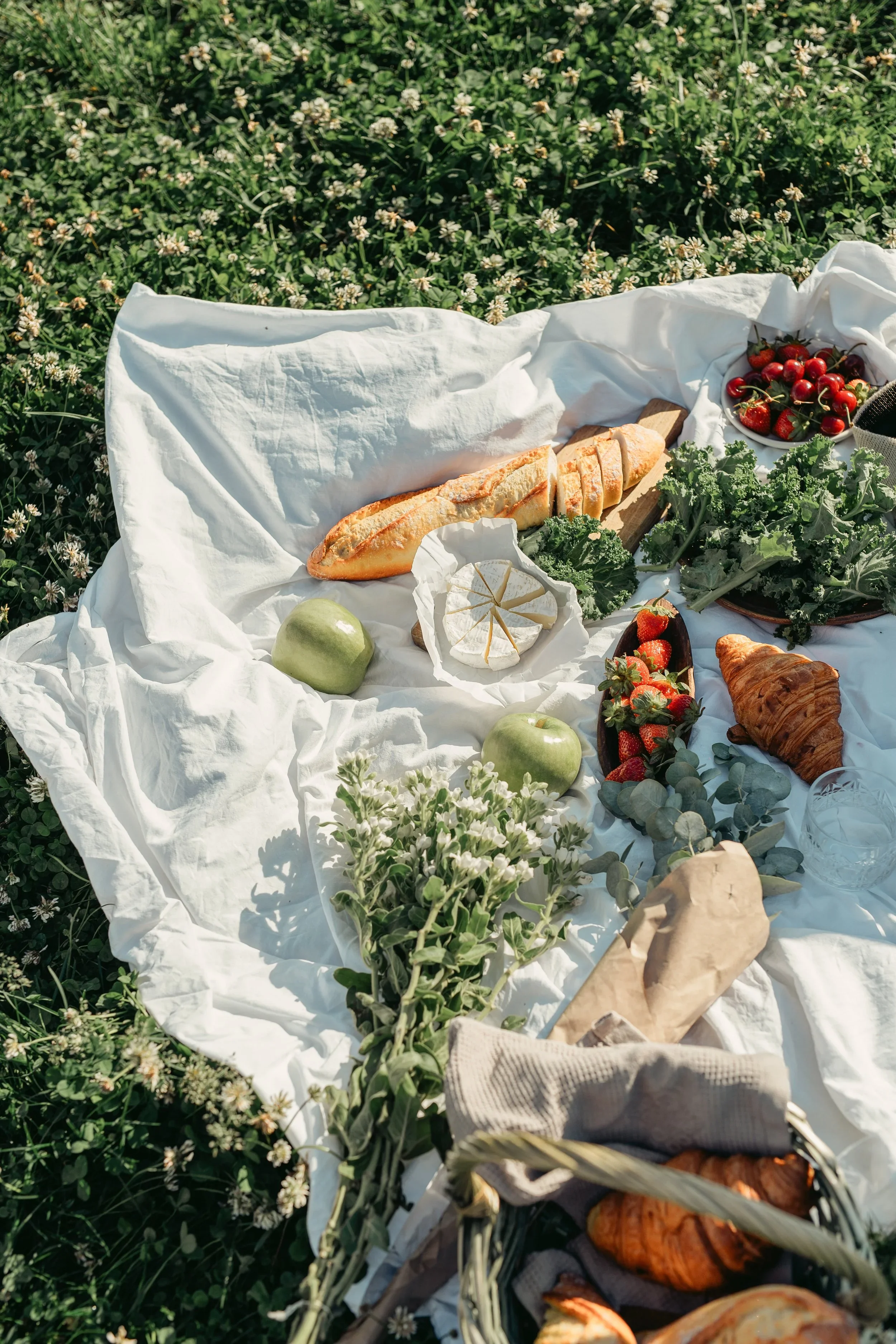 A picnic setup on white fabric covering grass, featuring baguette, green apples, strawberries, cherries, croissant, cheese, leafy greens, flowers, and glassware.