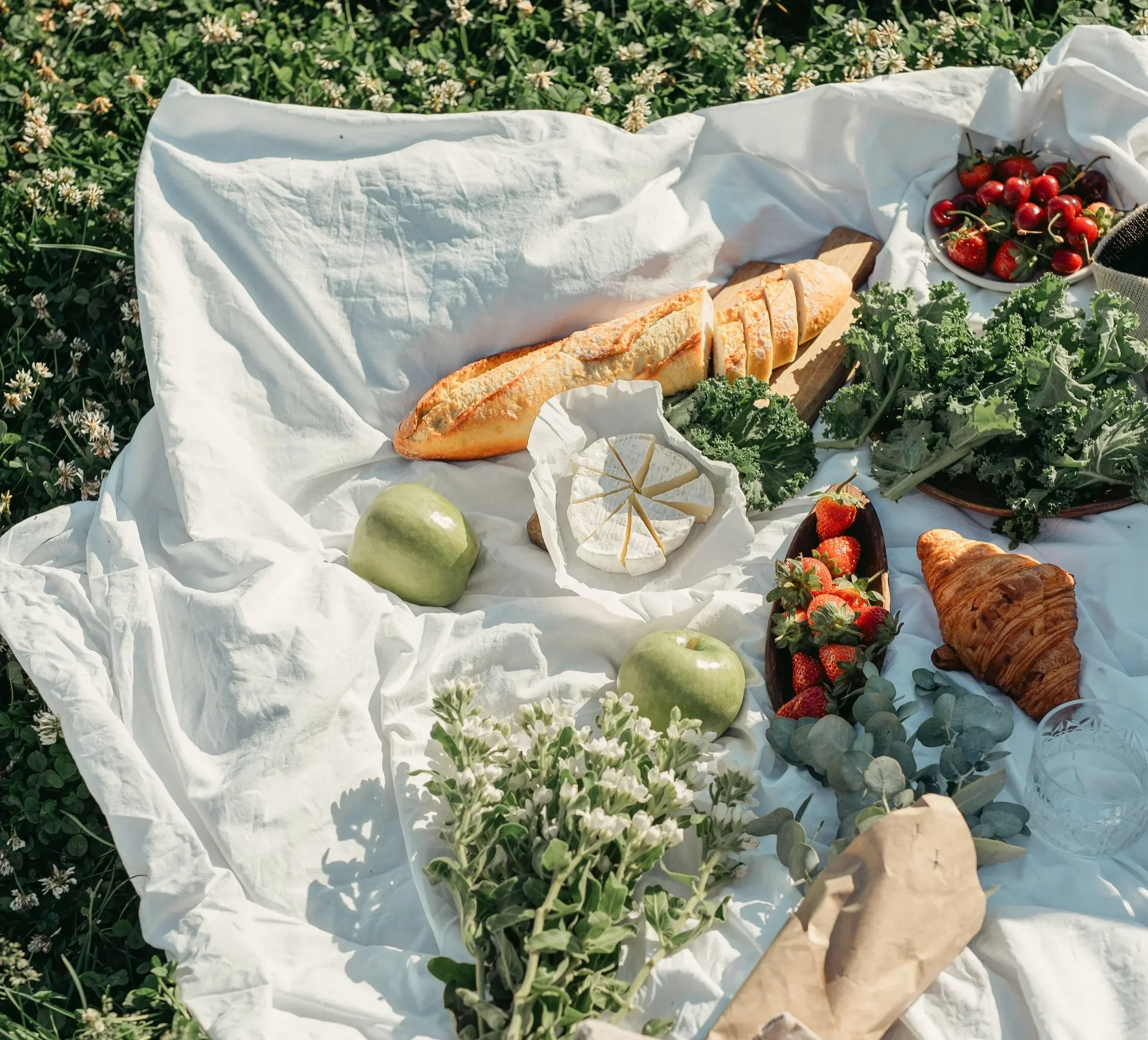A picnic setup with a white cloth spread on grass, featuring a baguette, a wheel of cheese, green apples, a croissant, strawberries, cherries, leafy greens, and a glass of water.