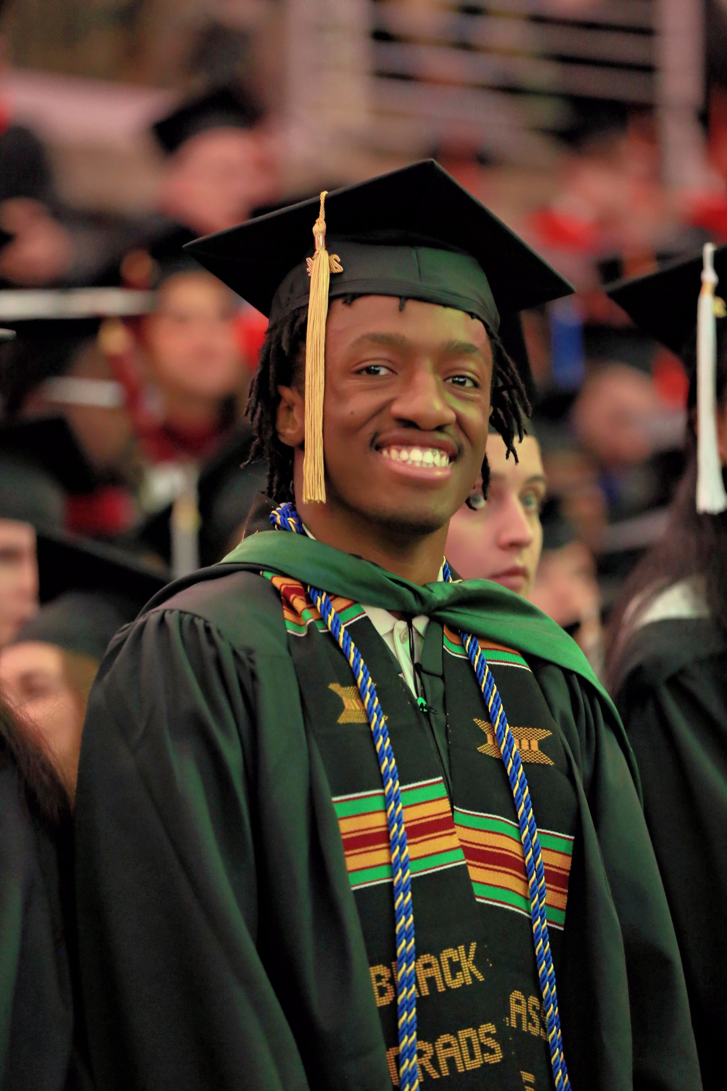 Young man in graduation cap and gown smiling during graduation ceremony, surrounded by other graduates in cap and gown.