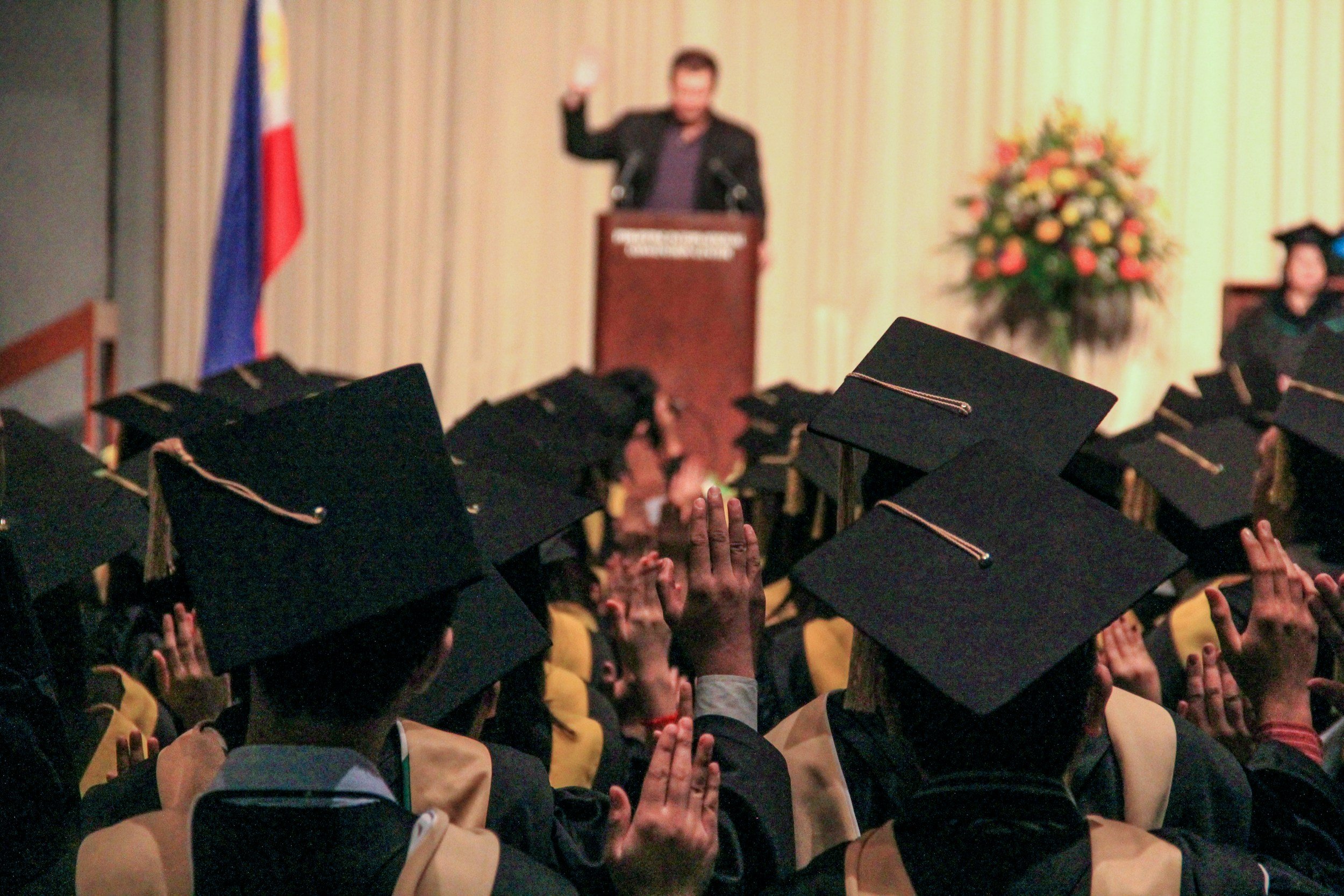 Graduates in caps and gowns raising their hands during a ceremony in a hall with a speaker at a podium in the background.