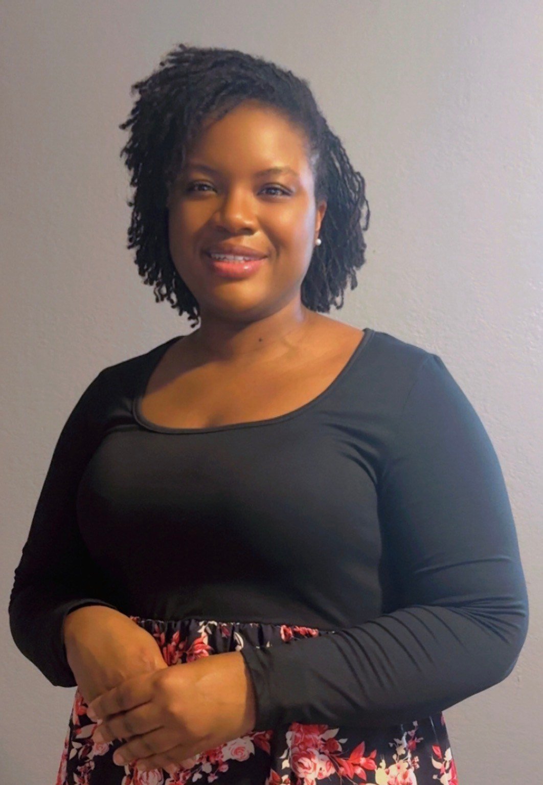 A woman with medium-dark skin and black curly hair styled in twists, wearing a black long-sleeve top and a floral skirt, standing against a plain light gray wall, smiling gently.