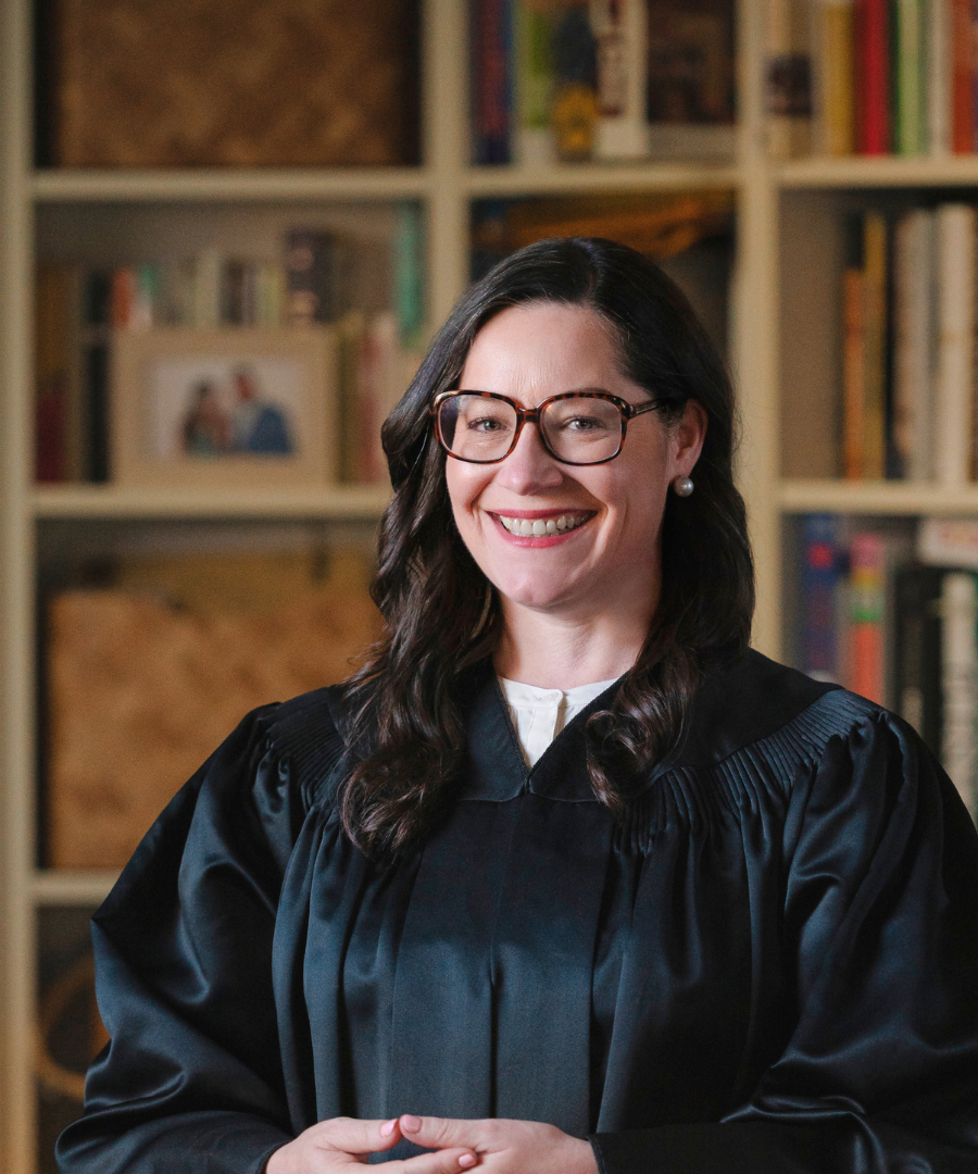 A woman in a black graduation gown smiling in front of a bookshelf.