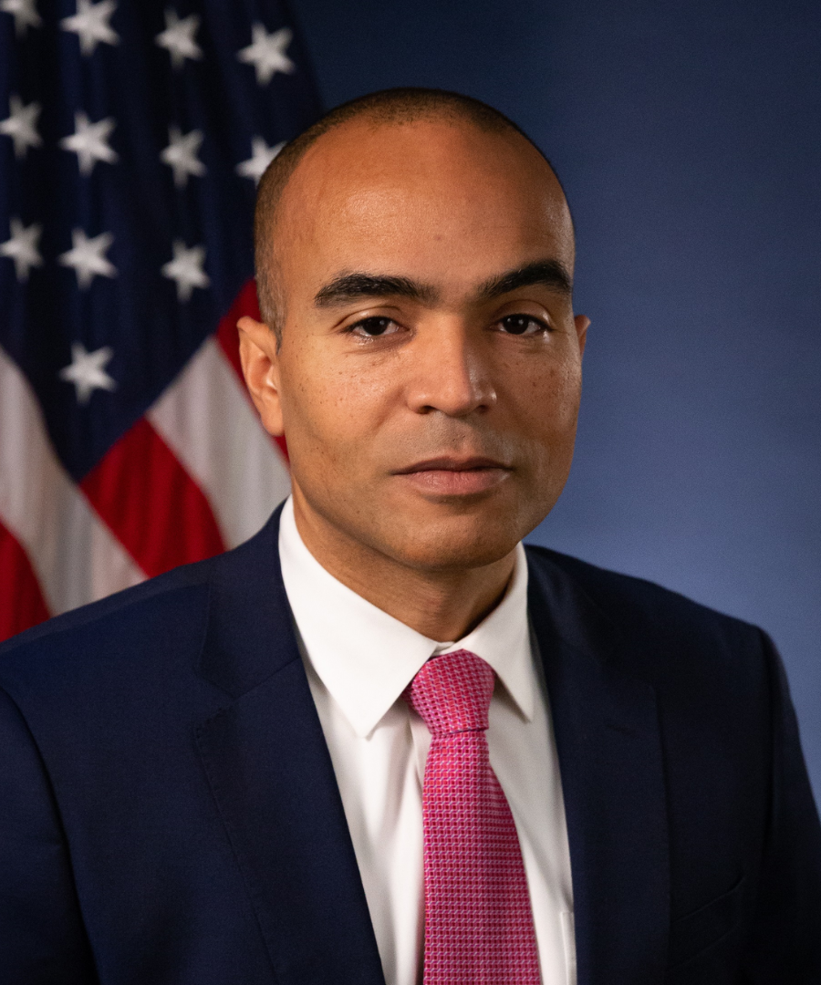 A man in a dark suit, white shirt, pink tie, standing in front of an American flag.