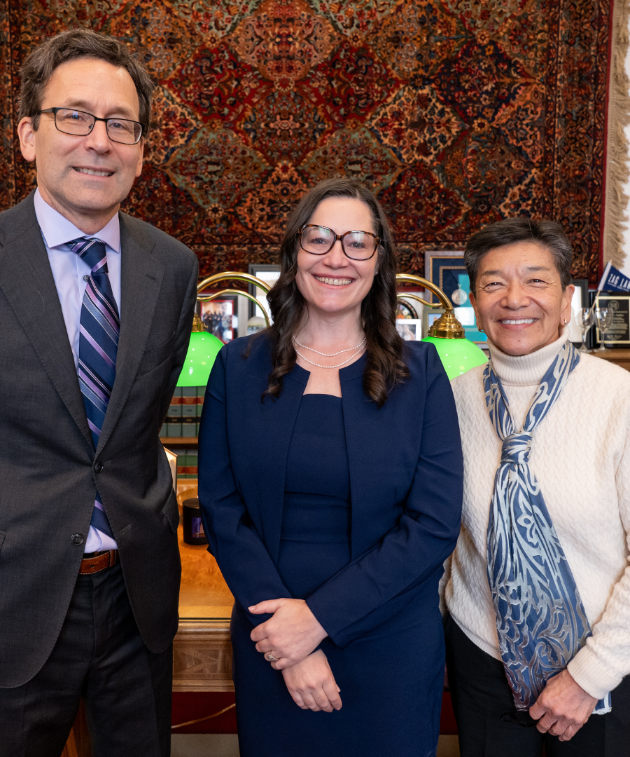 Three professionally dressed individuals standing in front of a decorative curtain with a desk behind them, smiling at the camera.