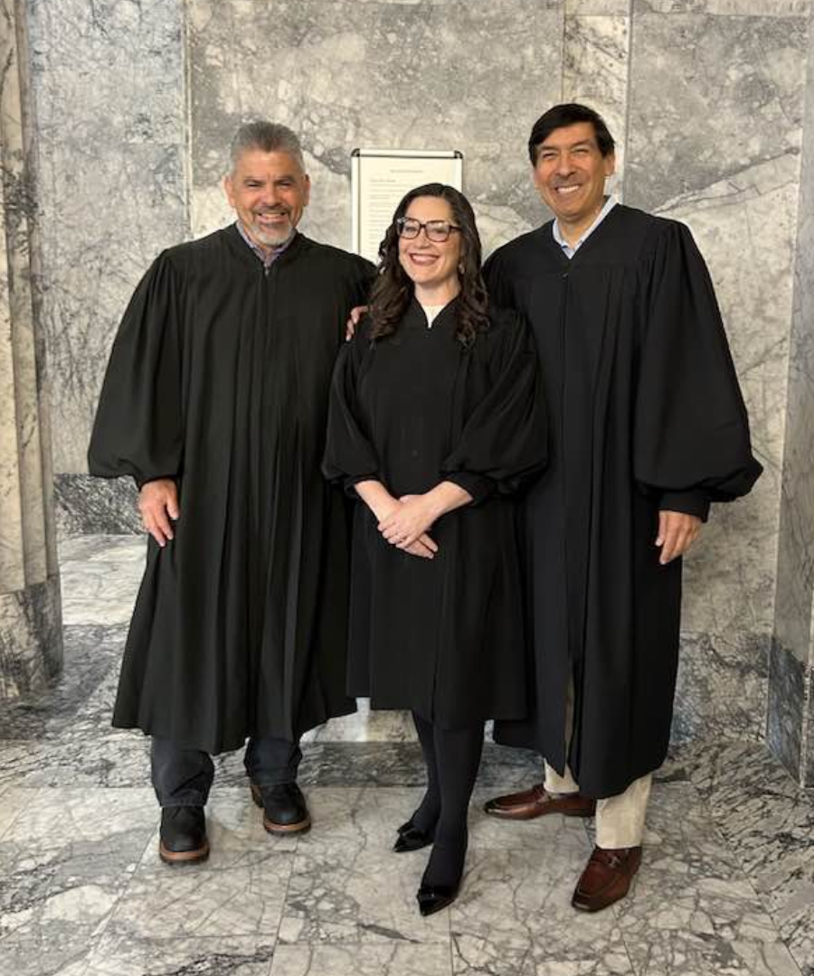 Three individuals in graduation gowns standing indoors in front of a marble wall, smiling at the camera.
