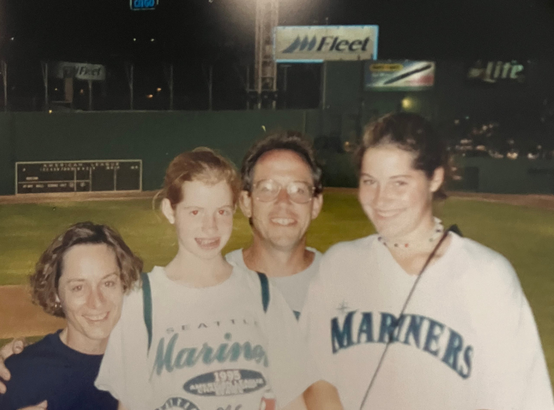 Four people standing together, two women and a man in the middle, in front of a baseball stadium at night. They are smiling, and some are wearing Seattle Mariners shirts.