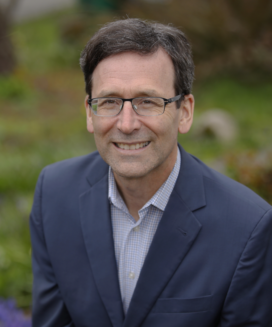 A middle-aged man with dark hair, glasses, and a friendly smile wearing a navy blazer and checkered shirt outdoors.