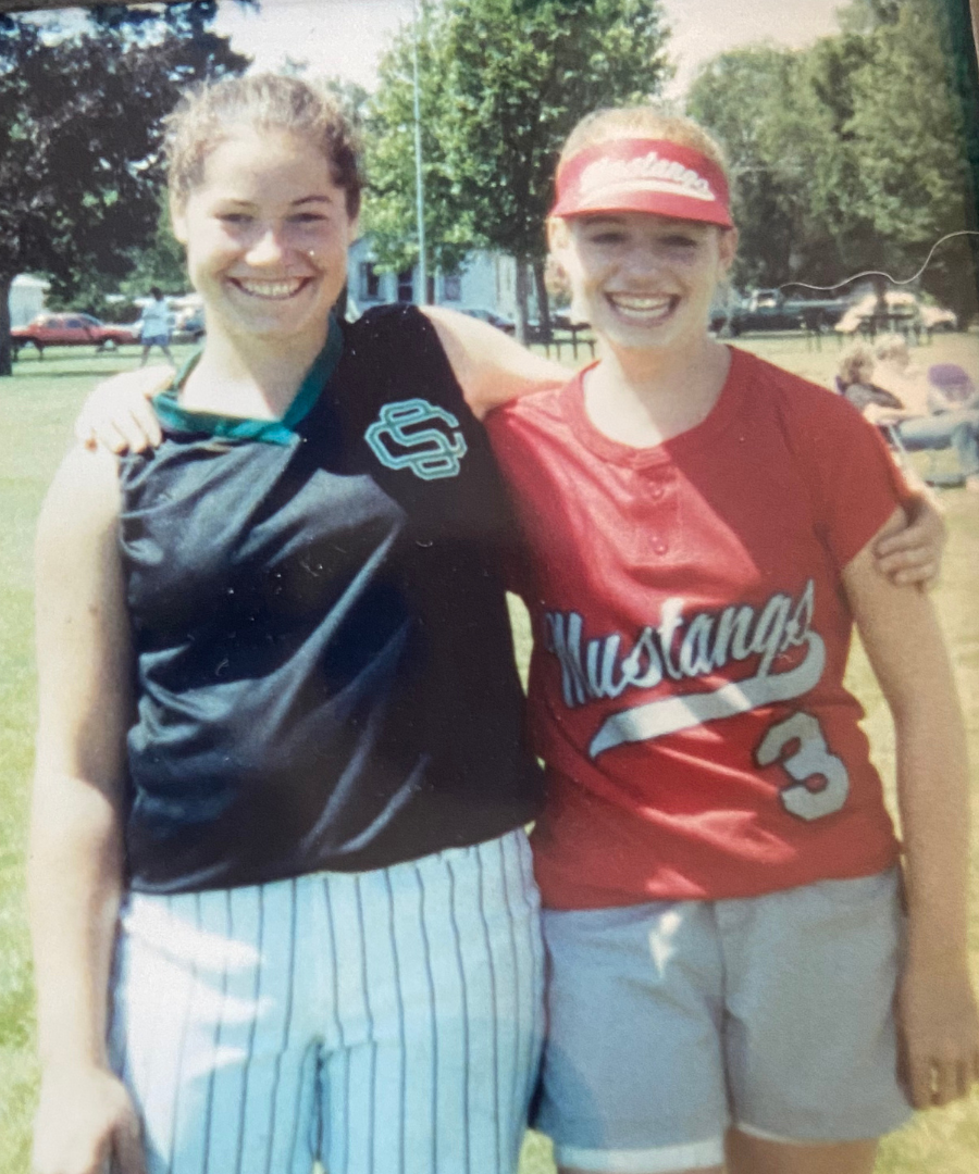 Two young women smiling and standing outdoors, with one wearing a black sports jersey with a logo and striped shorts, and the other wearing a red Mustang baseball jersey and shorts, with trees and parked cars in the background.