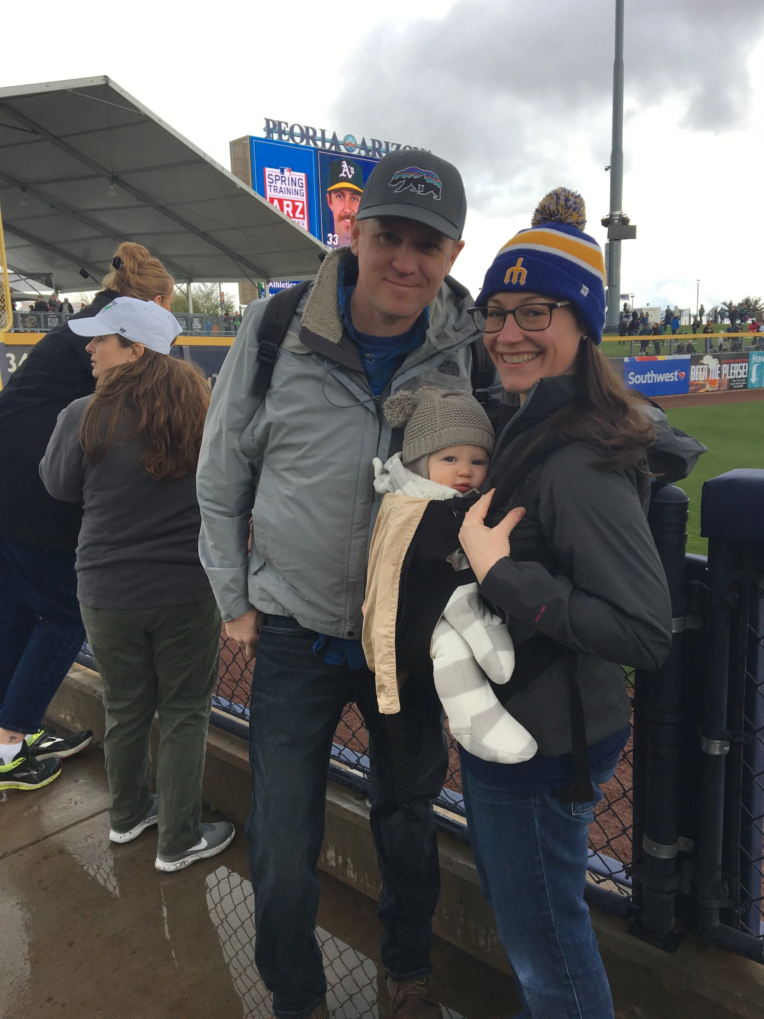Family at a baseball game, with a man, woman, and baby in the foreground, and a baseball field and scoreboard in the background.