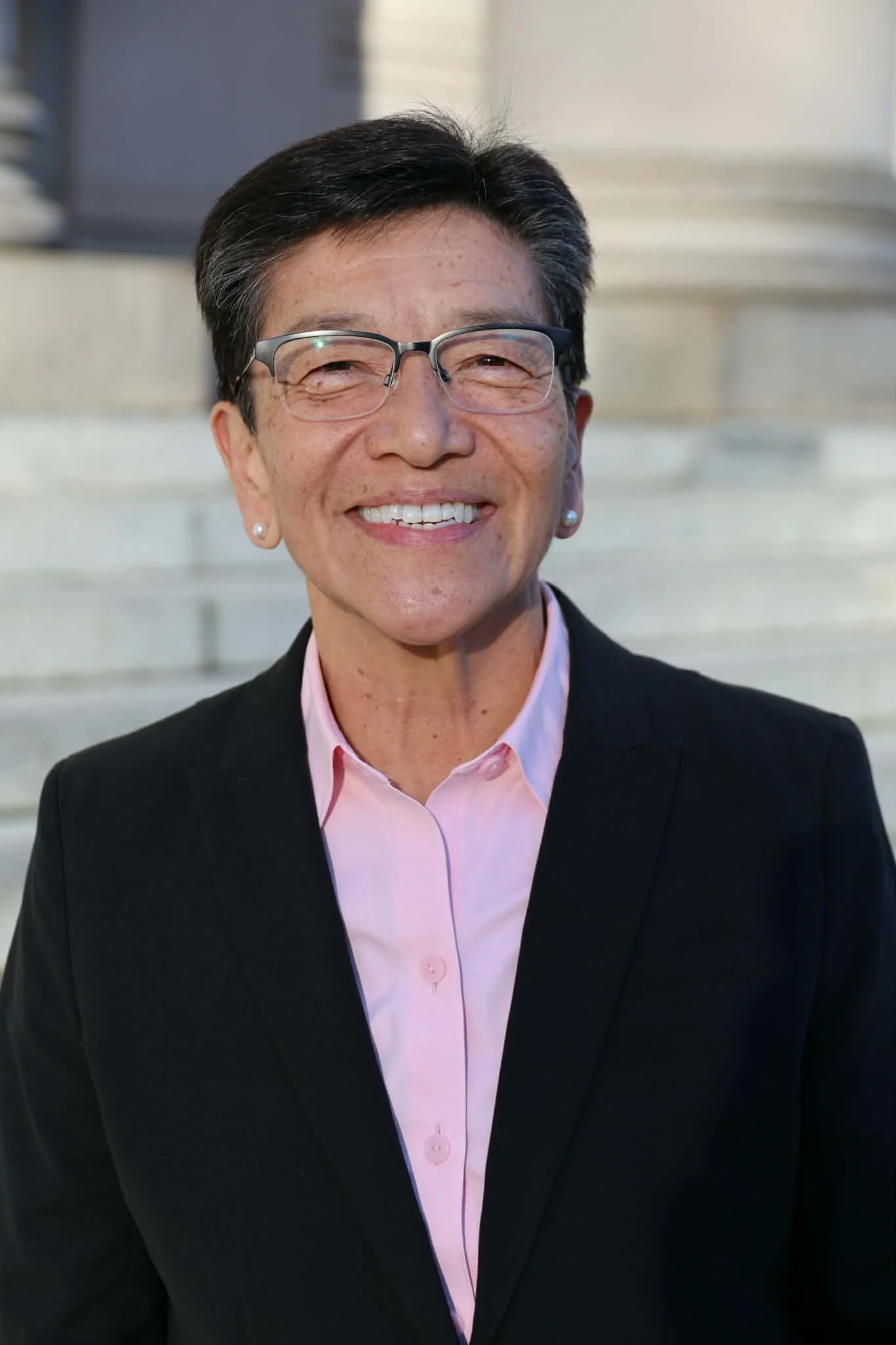 A woman with short black hair, smiling, wearing a dark navy blazer, earrings, and a necklace.
