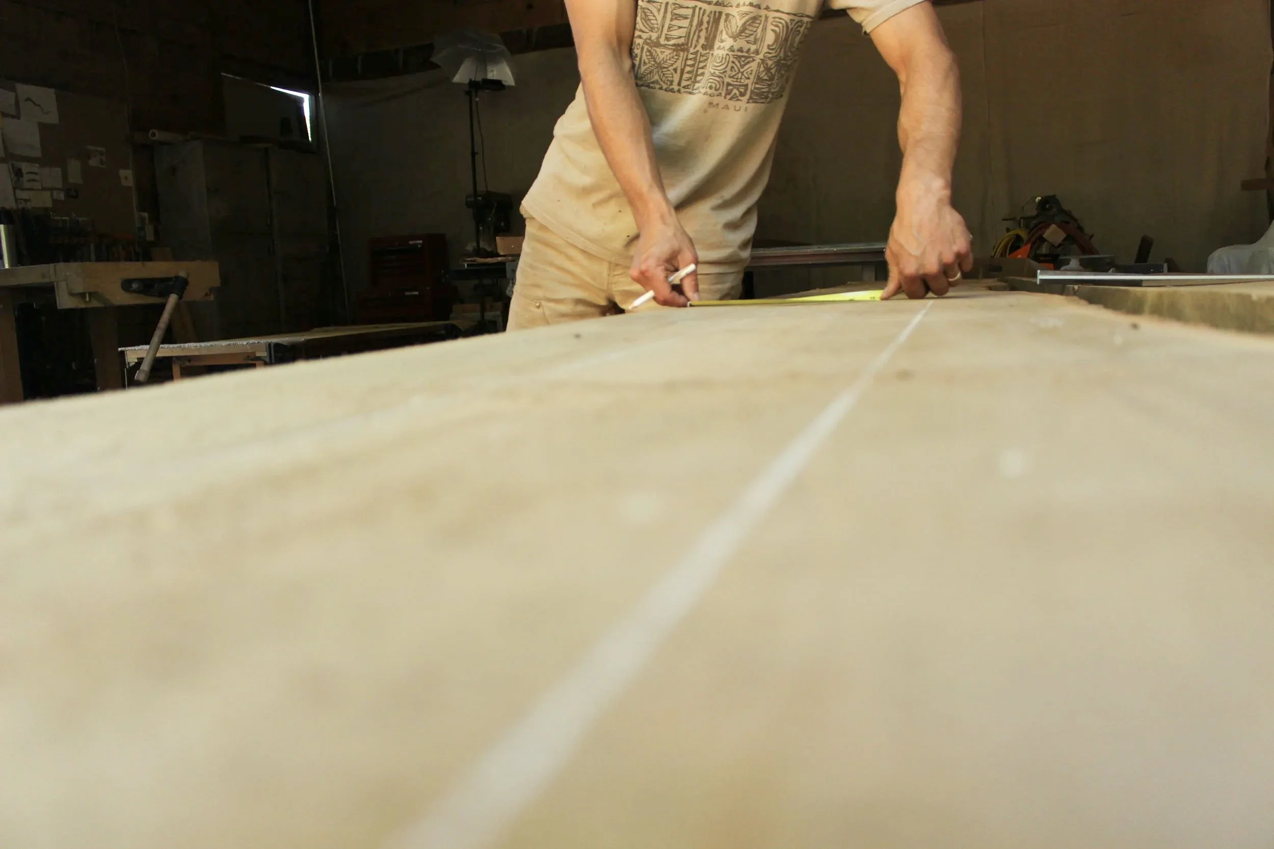 A person measuring a wooden board in a workshop with tools and equipment in the background.