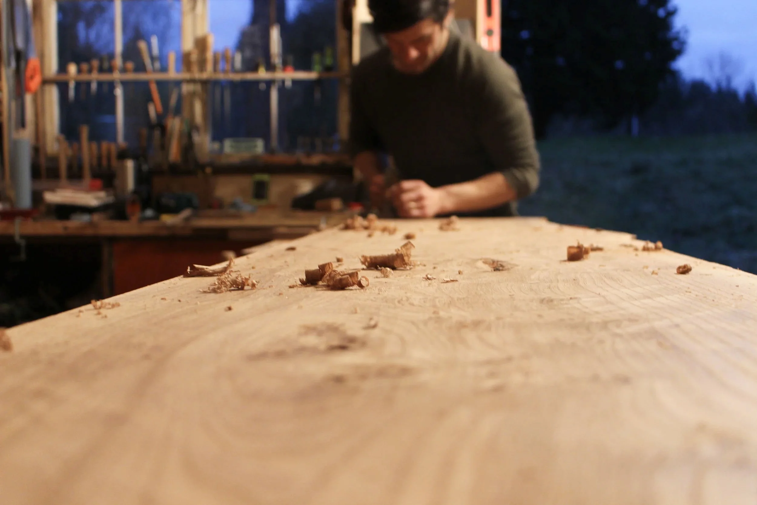 A man working on a wooden workbench outdoors during dusk, with wood shavings scattered on a large wooden surface in the foreground and a blurry background of trees and workshop tools.