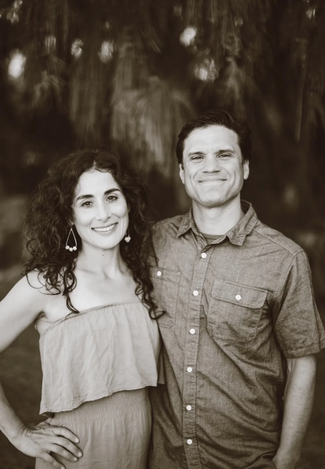A black and white photo of a smiling woman with curly hair, wearing a strapless dress, and a man with short hair in a button-up shirt, standing closely outdoors with blurred trees in the background.