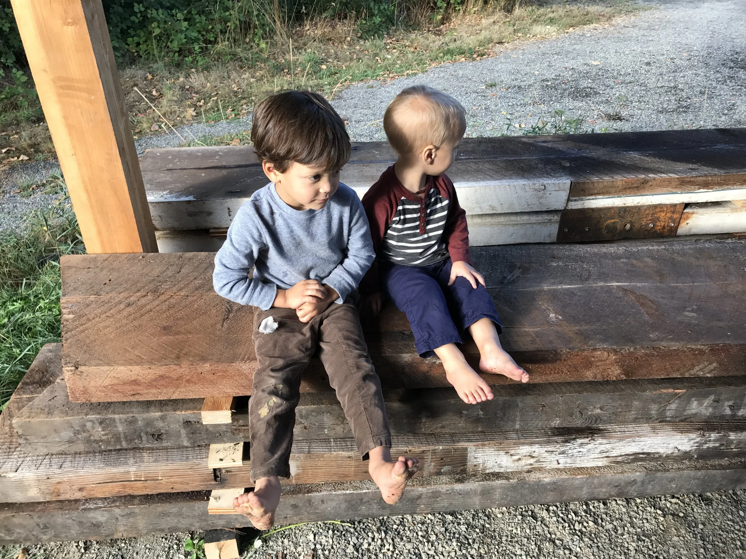 Two young boys sitting barefoot on a wooden structure outdoors, looking away from the camera. One boy has brown hair and is wearing a gray long-sleeve shirt and brown pants, while the other boy has blonde hair and is wearing a maroon and striped shirt with blue pants.