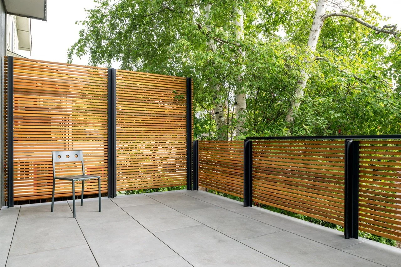 Empty balcony with a single gray chair, wooden slat privacy screen, gray tiled floor, and lush green trees in the background.