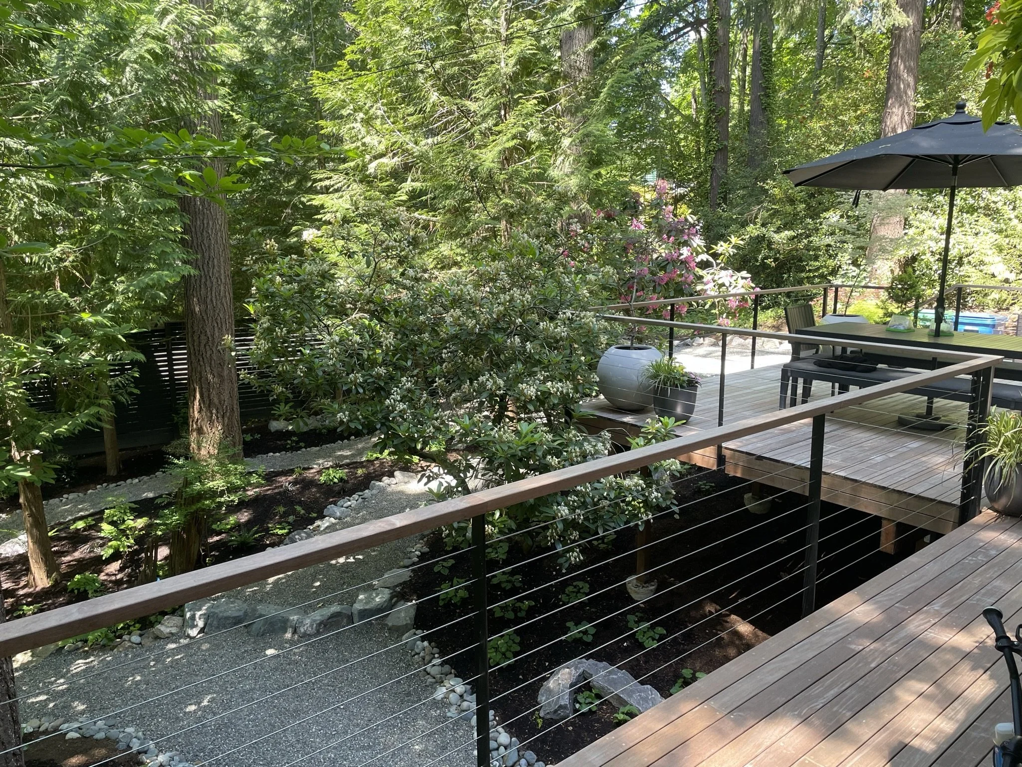 A wooden deck with outdoor furniture and large potted plants, overlooking a lush green garden with trees and flowering bushes, in a peaceful backyard setting.