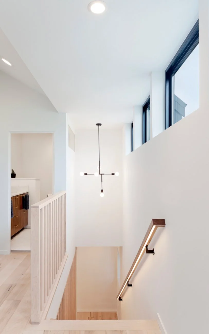 Interior view of a modern home staircase with white walls, black-framed windows, a wooden handrail, and a minimalist black chandelier hanging from the ceiling.